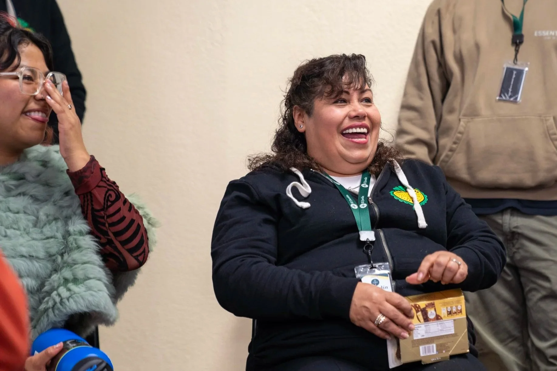Two people laughing and smiling indoors, one holding a snack package, and another wearing glasses and a fluffy jacket.