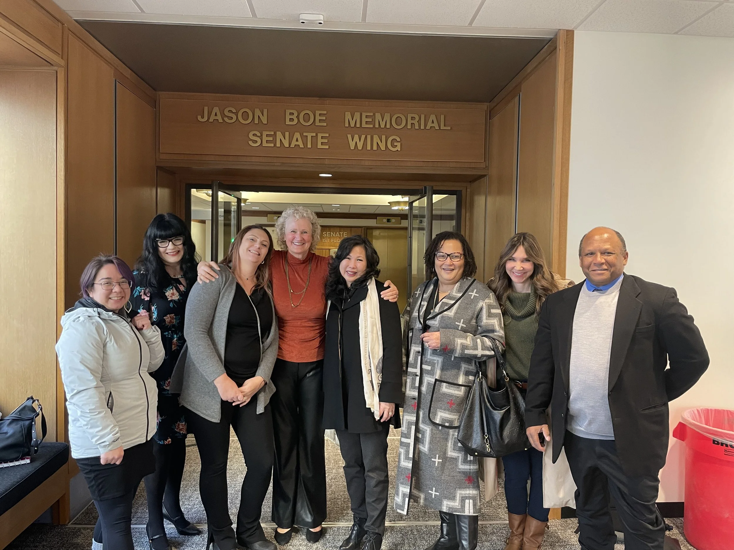 A group of eight HJRA advocates together in front of a sign that reads 'Jason Boe Memorial Senate Wing', smiling and posing for a photo inside a building.