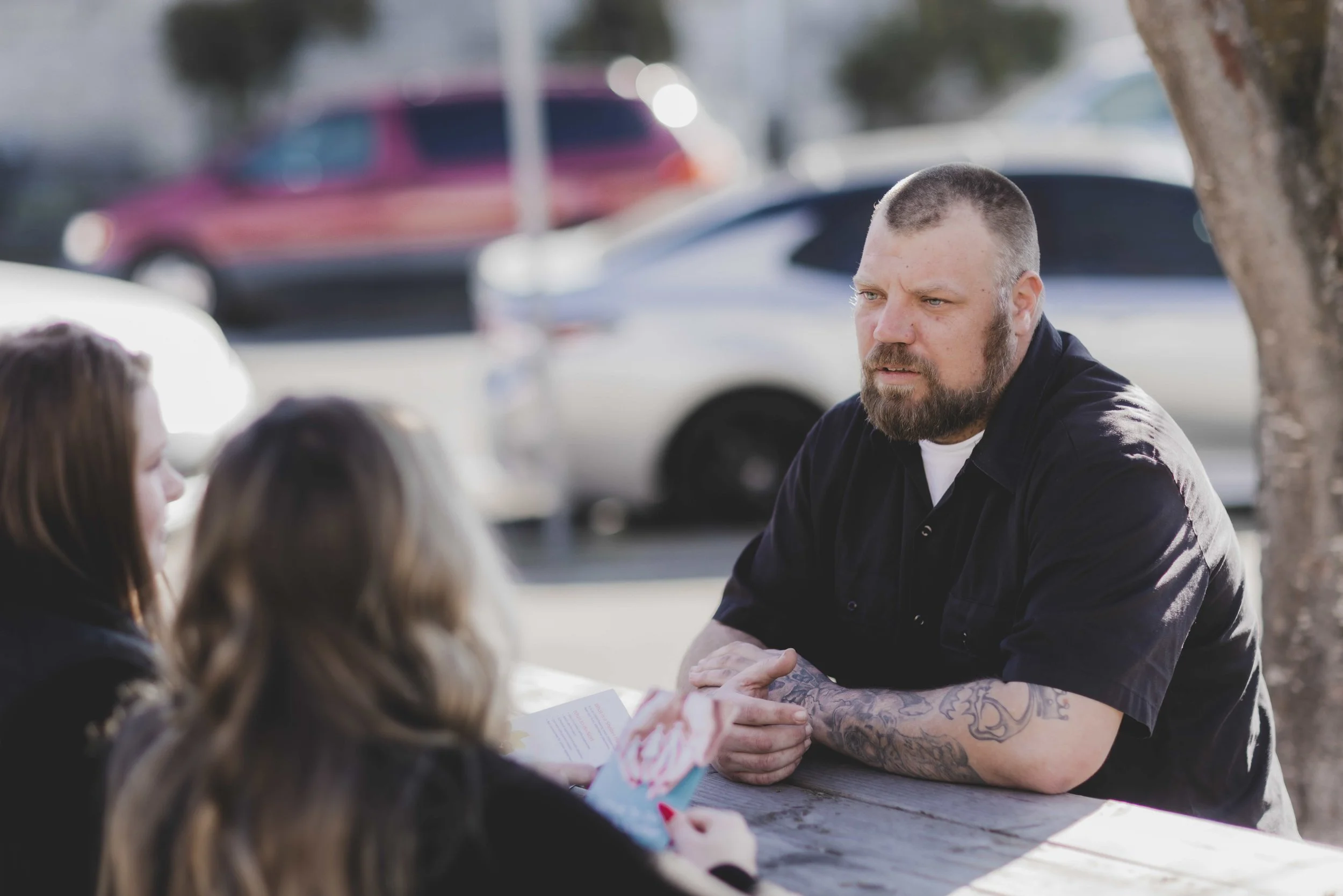 A man with a beard and tattoos on his arms part of Family Nurturing Center sits at a picnic table, talking to two women in an outdoor setting with cars in the background.