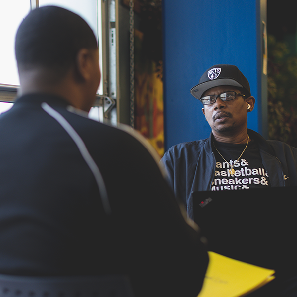 Two Black men from Fresh Out have a conversation in an indoor setting, with one facing away from the camera and the other wearing a baseball cap and glasses, sitting at a table with a yellow notepad.