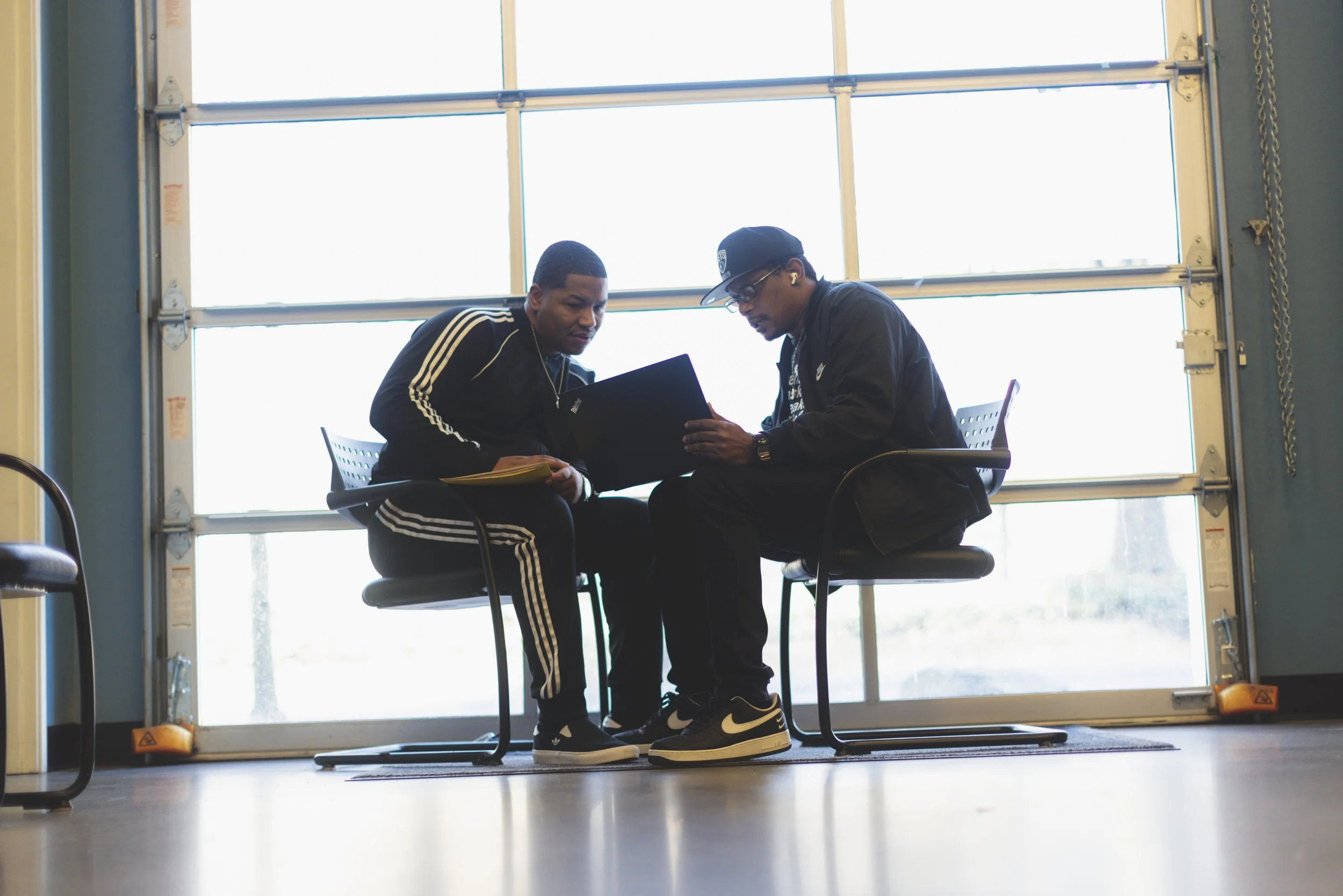 Two young Black men from Fresh out sit in front of a large glass window, looking at a laptop and talking.