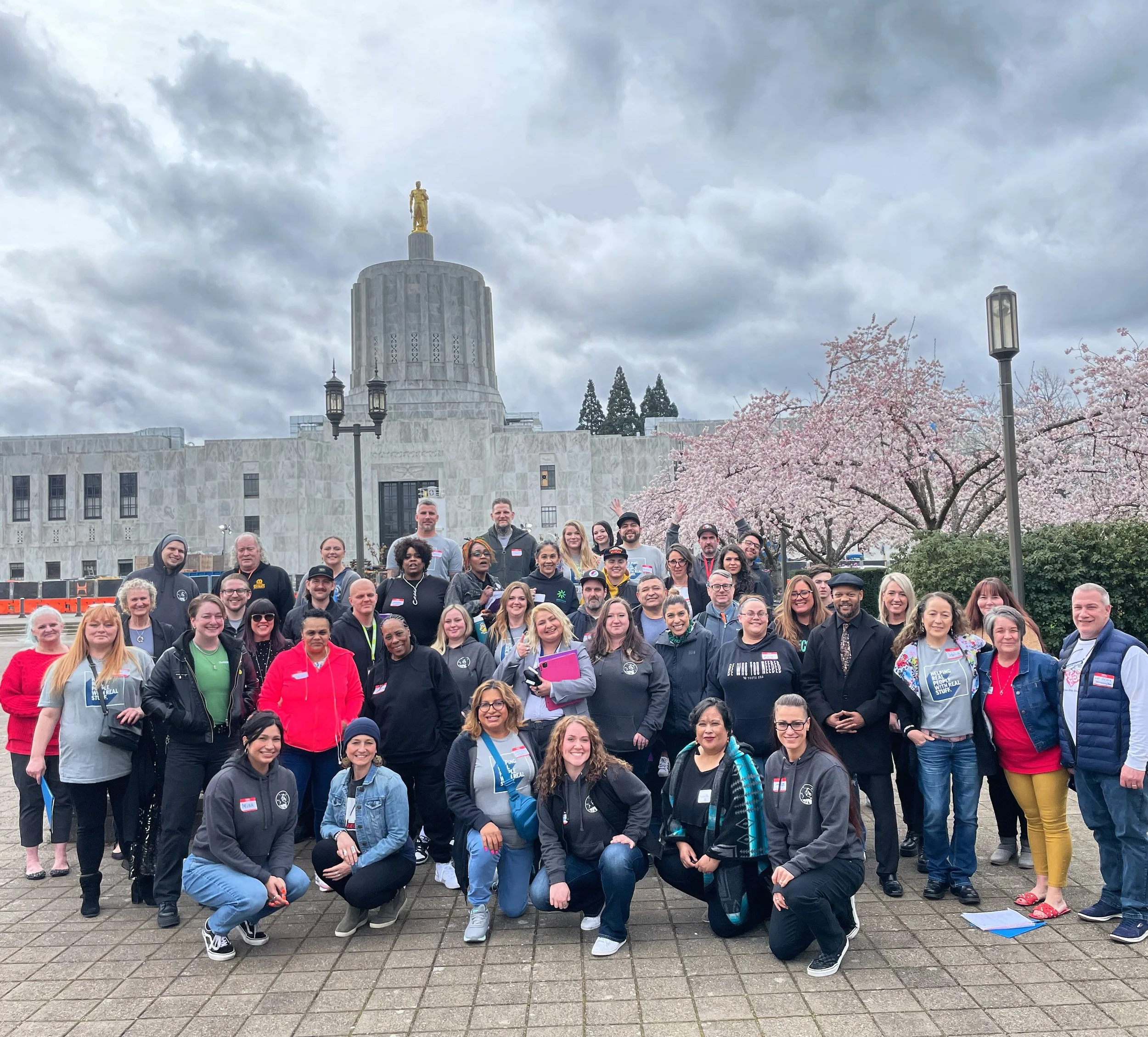 A group of diverse advocates pose outside the state capital, surrounded by cherry blossom trees.