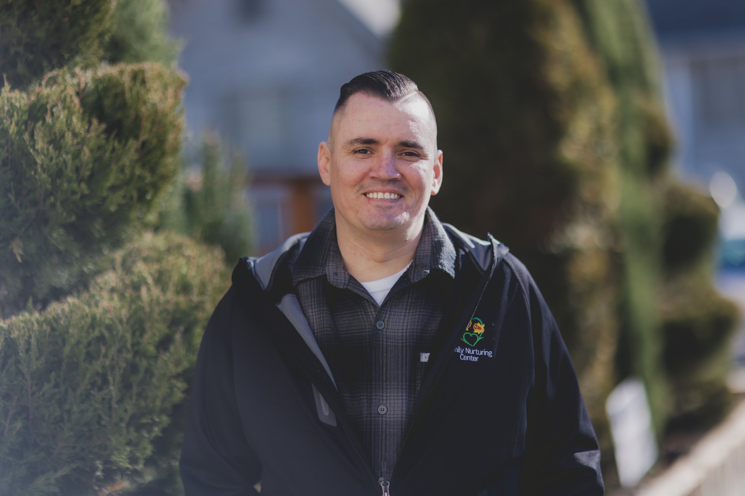 A provider at Miracles Club smiles, wearing a dark jacket and plaid shirt and standing outdoors with trees and bushes in the background.
