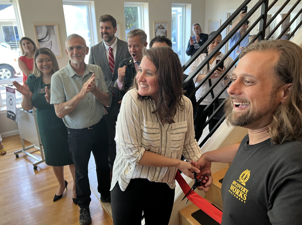 Two people cut a ribbon while others smile and clap behind them at the grand opening of Recovery Works.
