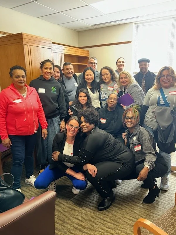Group of HJRA advocates pose for a group photo, some wearing casual clothing and name tags, with wooden cabinets and a large window in the background 
