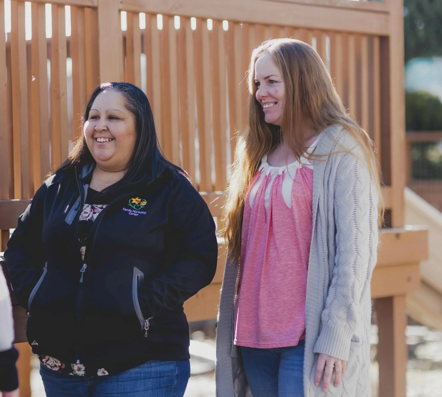 Two people, both with loose, long hair, smile together, one wearing a branded fleece and the other in a pink top and gray sweater. They are both from Family Nurturing Center, a Measure 110 grantee in Southern Oregon.