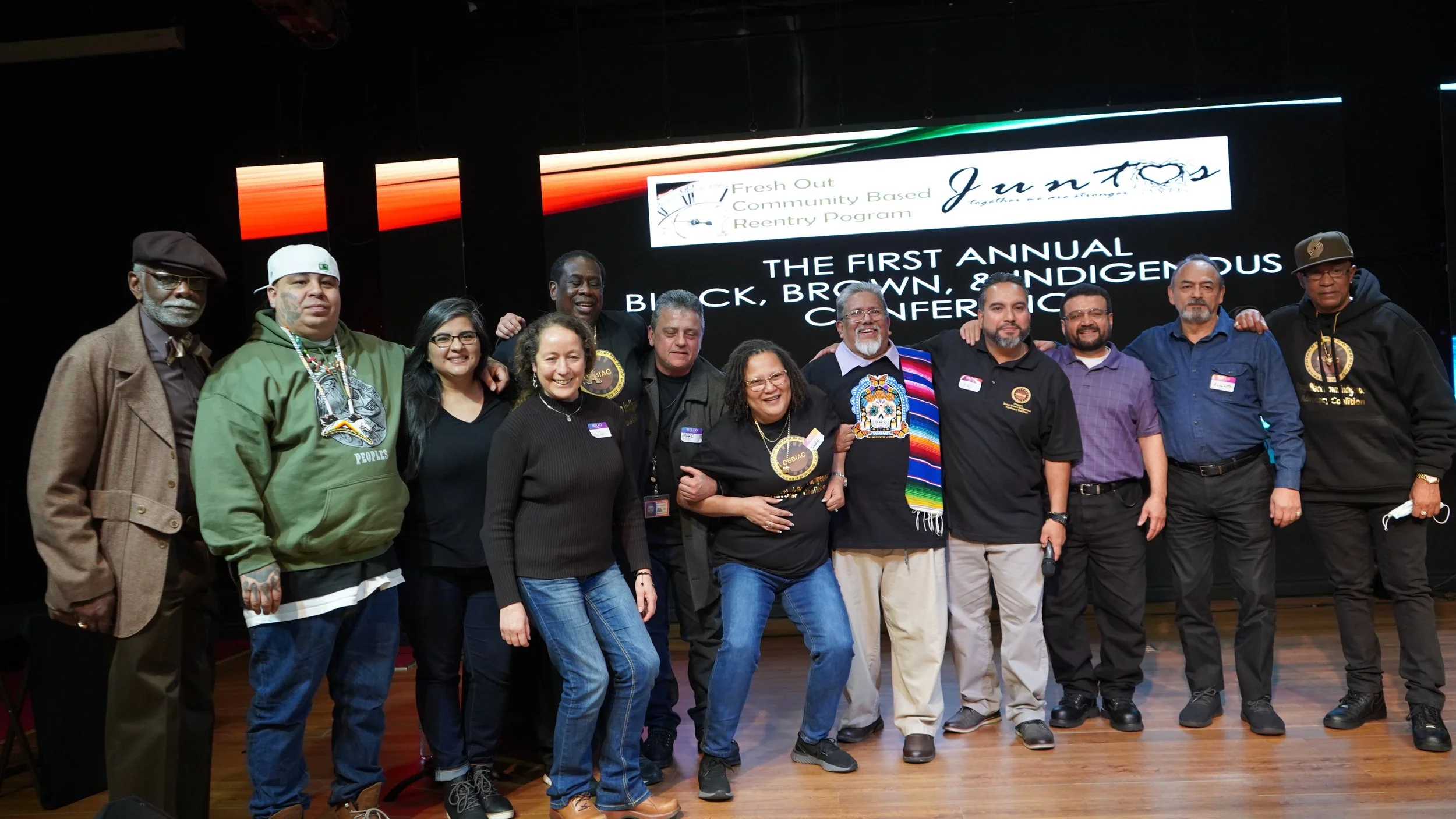 A group of people, mostly Indigenous, stand together on stage at the first Black Brown Indigenous Conference hosted by Fresh Out Community-Based Reentry centering culturally-specific recovery and reentry strategies.