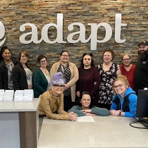 Team at Adapt stand and sitting in front of a brick wall with the word 'adapt' on it, in a reception area.