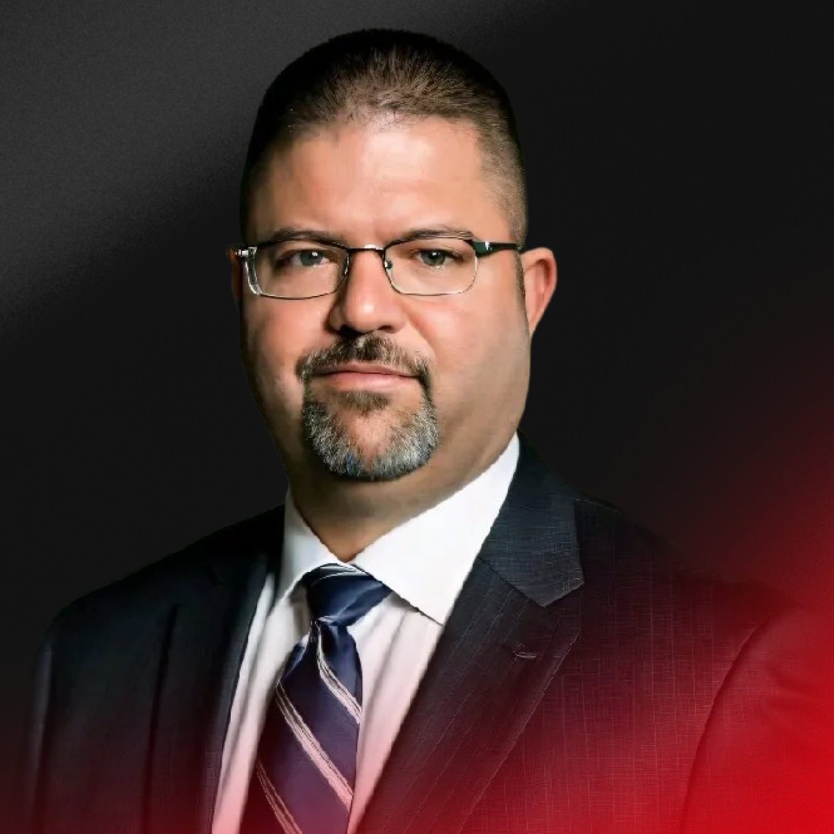A man with glasses, a goatee, and short hair in a business suit against a dark background.