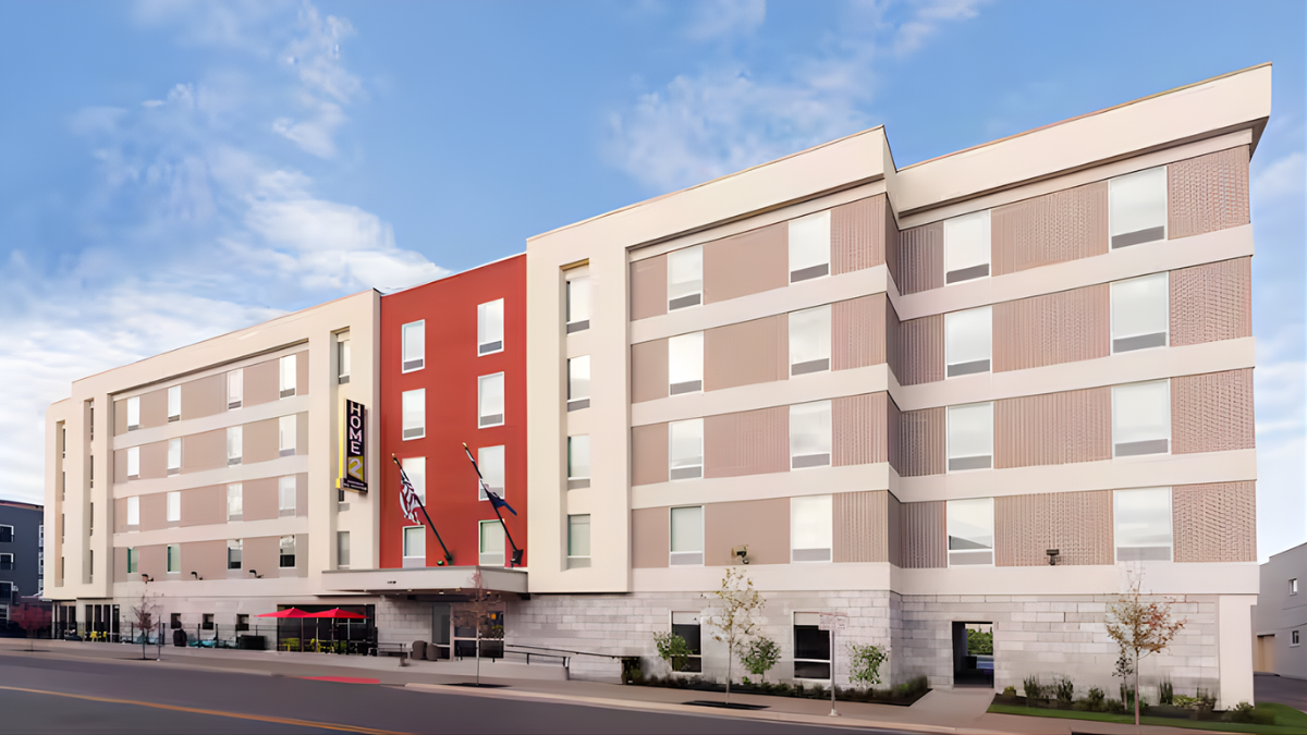 Multi-story modern hotel building with a red accent wall, flags near the entrance, storefront with umbrellas, and a parking area in front.