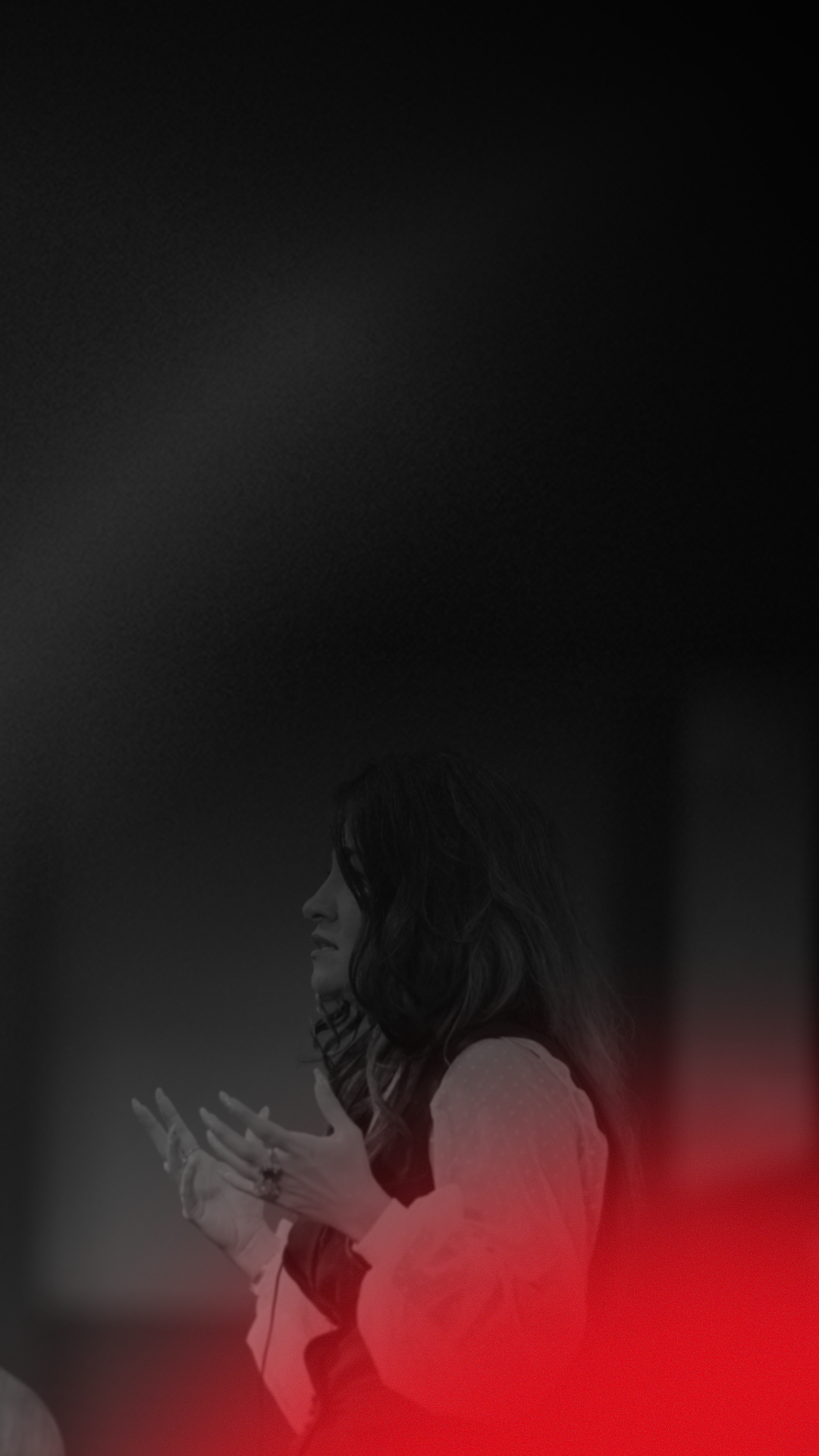 A woman with dark, wavy hair, wearing a light-colored, long-sleeved shirt, gestures with her hands in front of her during a presentation or speech, with a blurred background.