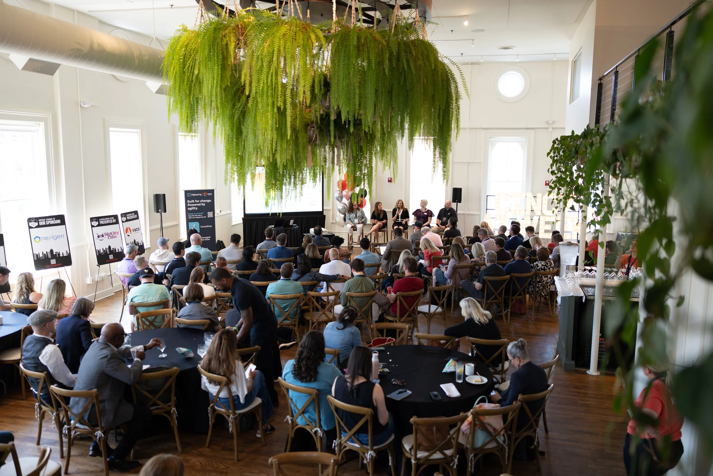 A large conference room filled with round tables and chairs, where a panel discussion is happening on stage with five speakers, and audience members listening. The room has a high ceiling with large windows and hanging green plants.