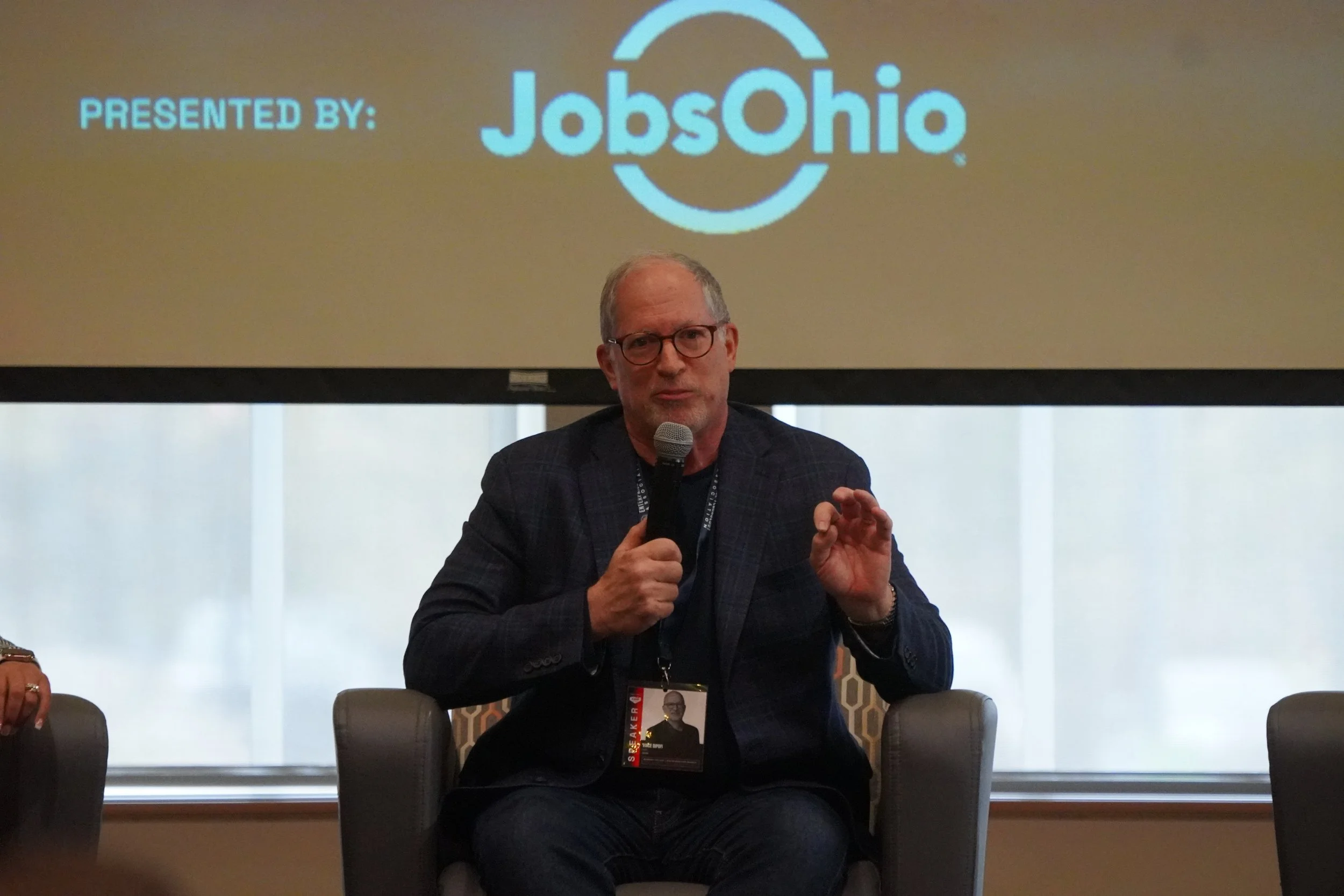 A man with glasses holding a microphone sits on a chair in front of a large screen displaying 'JobOhio' and 'Presented by:'.