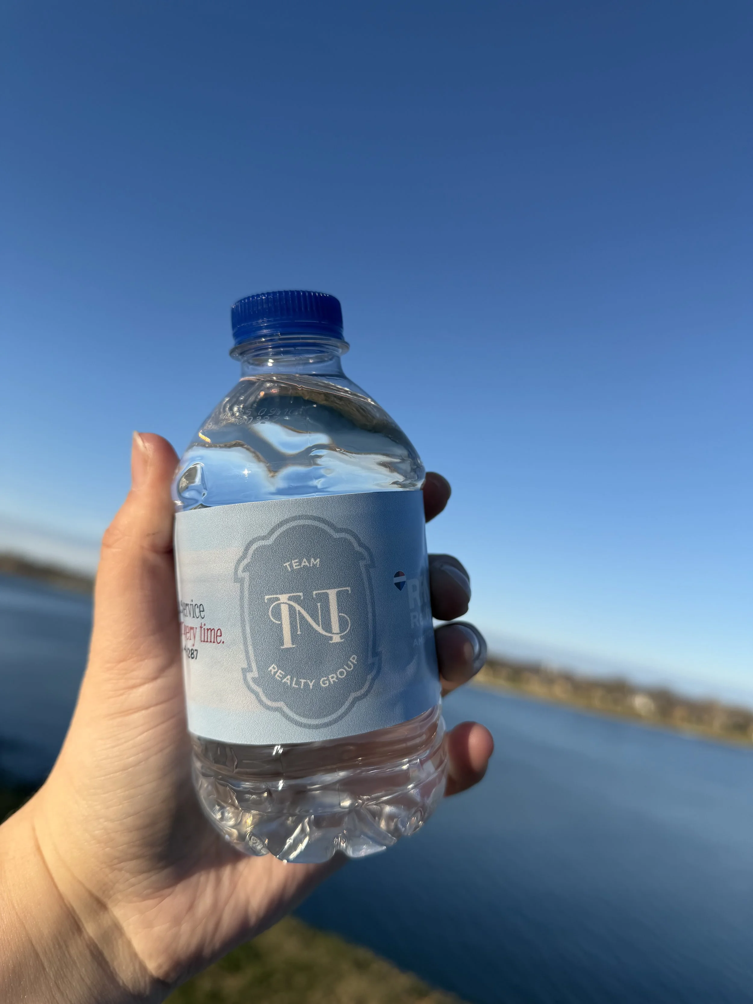 A person holding a bottle of water with a blue cap and a label that reads 'Team Realty Group' against a clear blue sky and a body of water in the background.