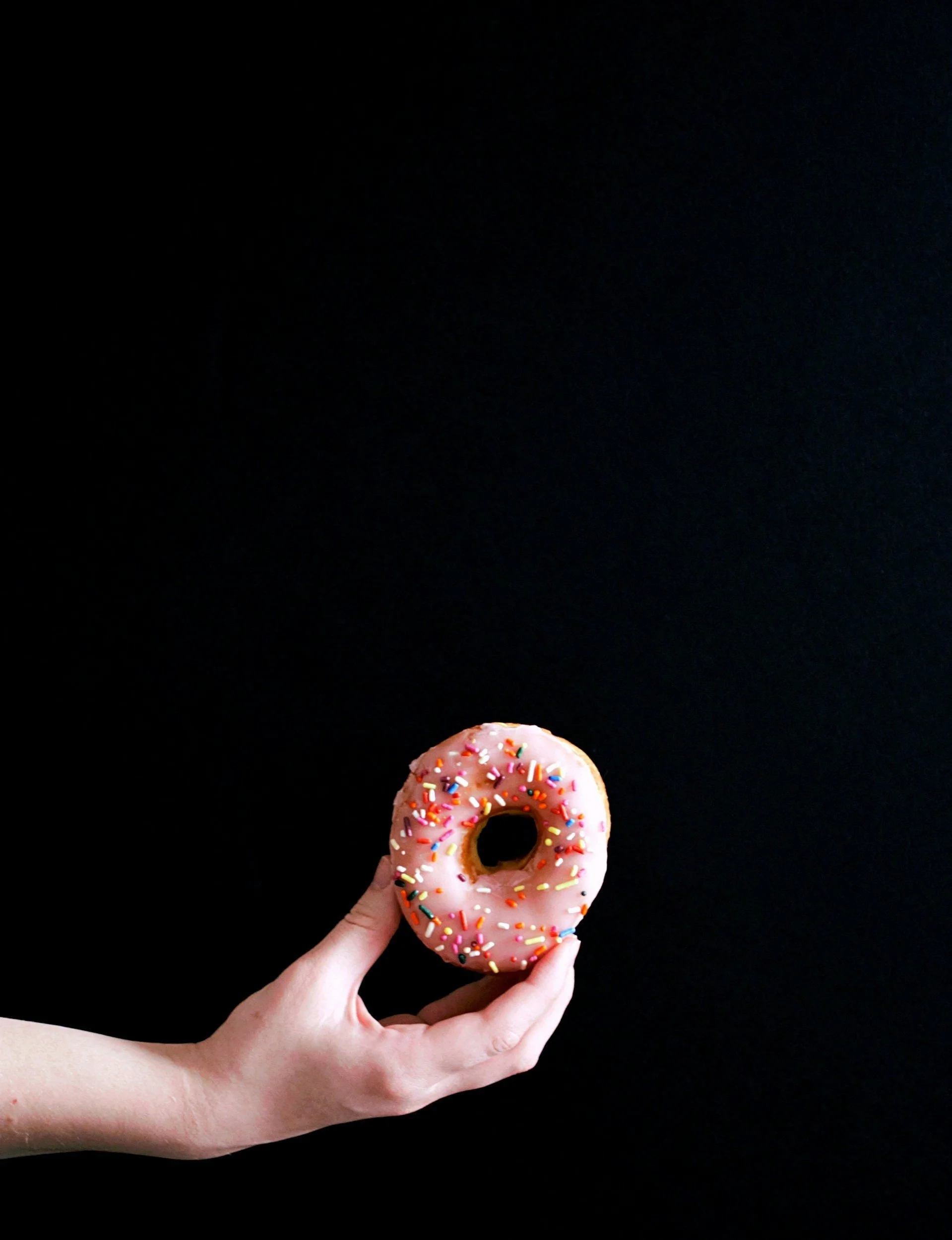 Hand holding a pink frosted donut with colorful sprinkles against a black background.