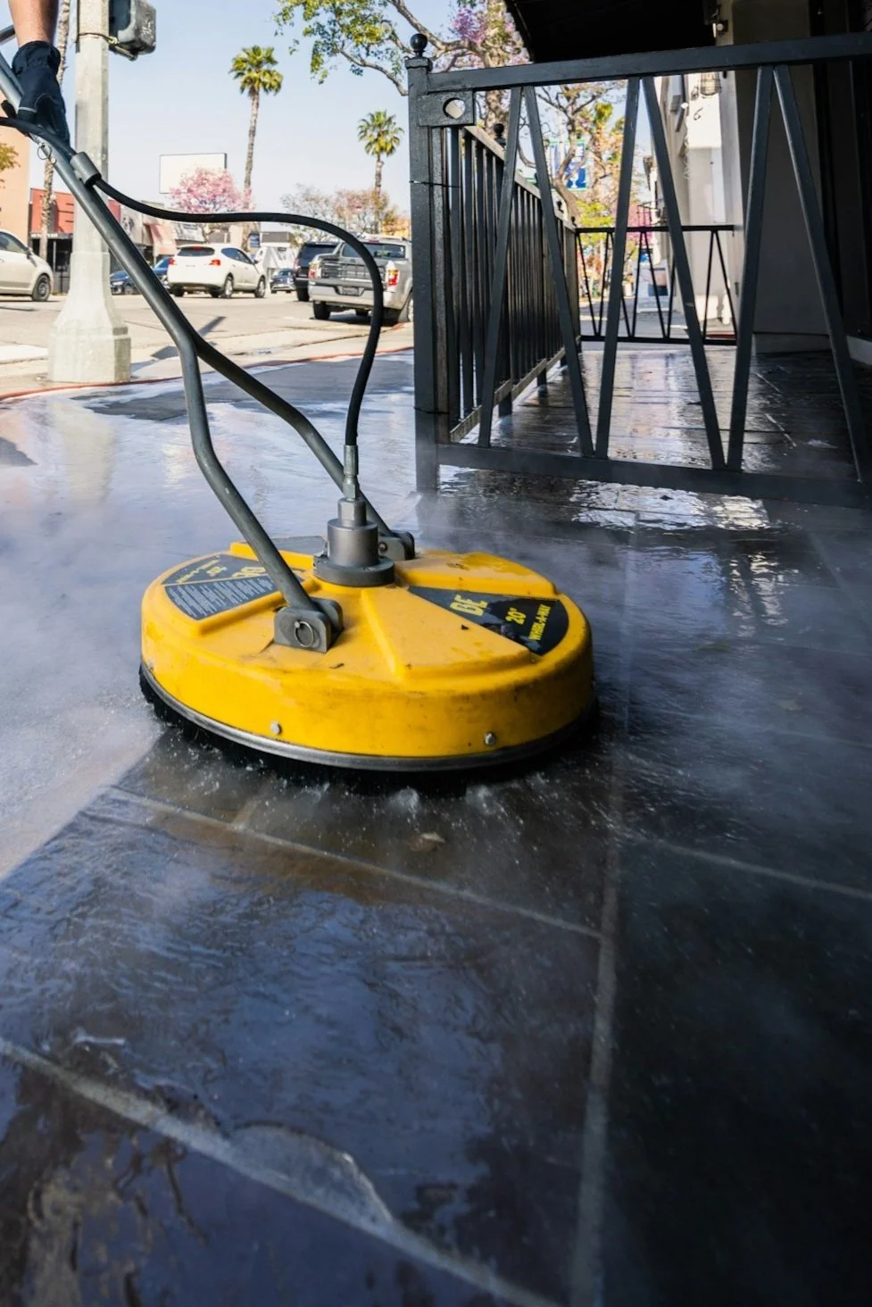 A yellow floor buffer in use on a dark tile floor outside a storefront, with parked cars, palm trees, and a basketball hoop in the background.