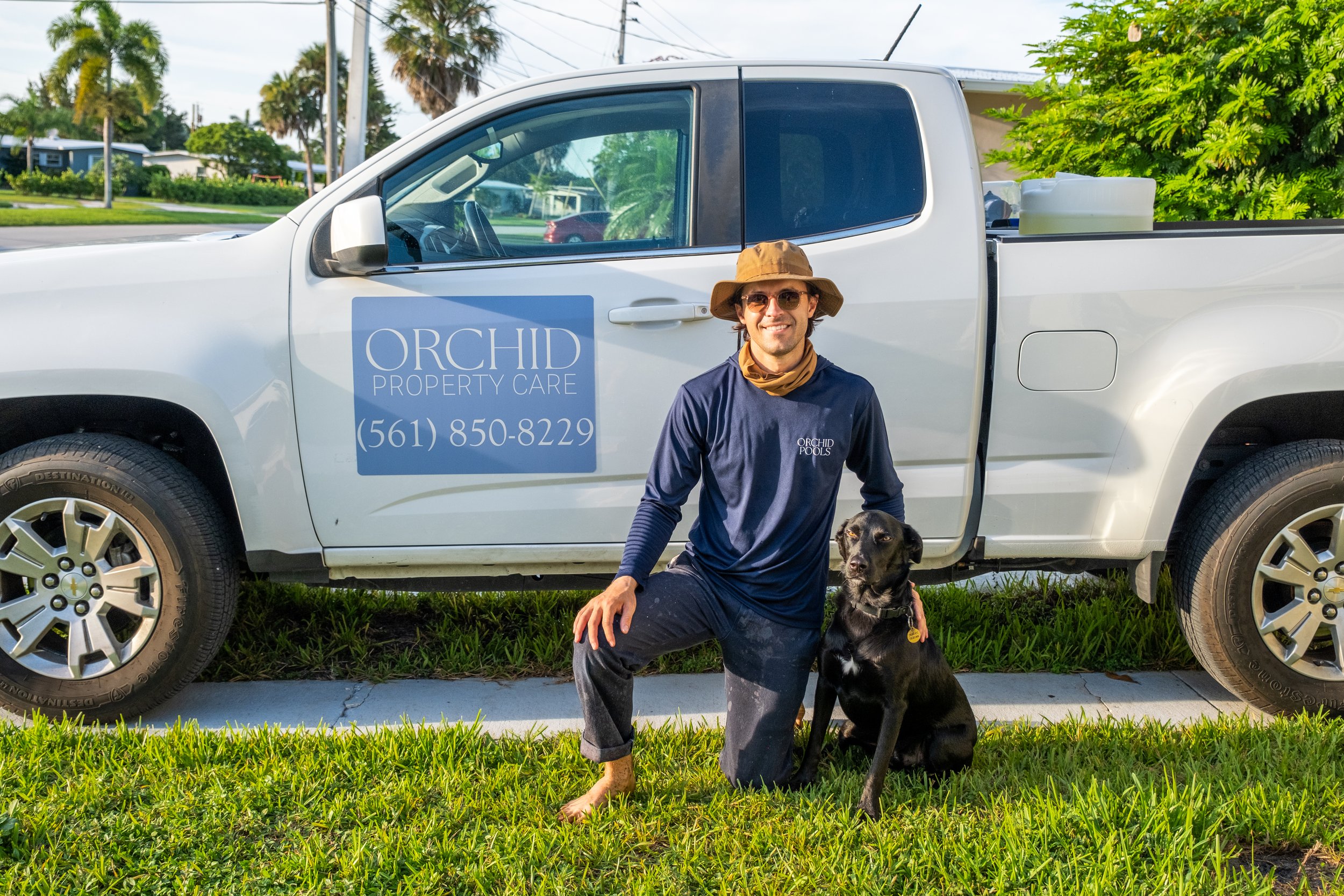Man in hat and sunglasses kneeling beside a black dog on a grass patch in front of a white pickup truck with 'Orchid Property Care' sign and phone number.