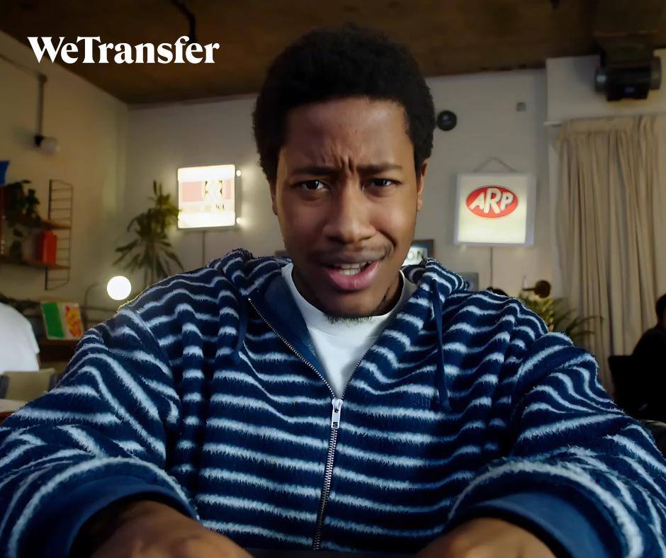A young man with a surprised or confused expression taking a selfie indoors, with a sign that reads 'WeTransfer' and another sign with 'ARP' in the background.