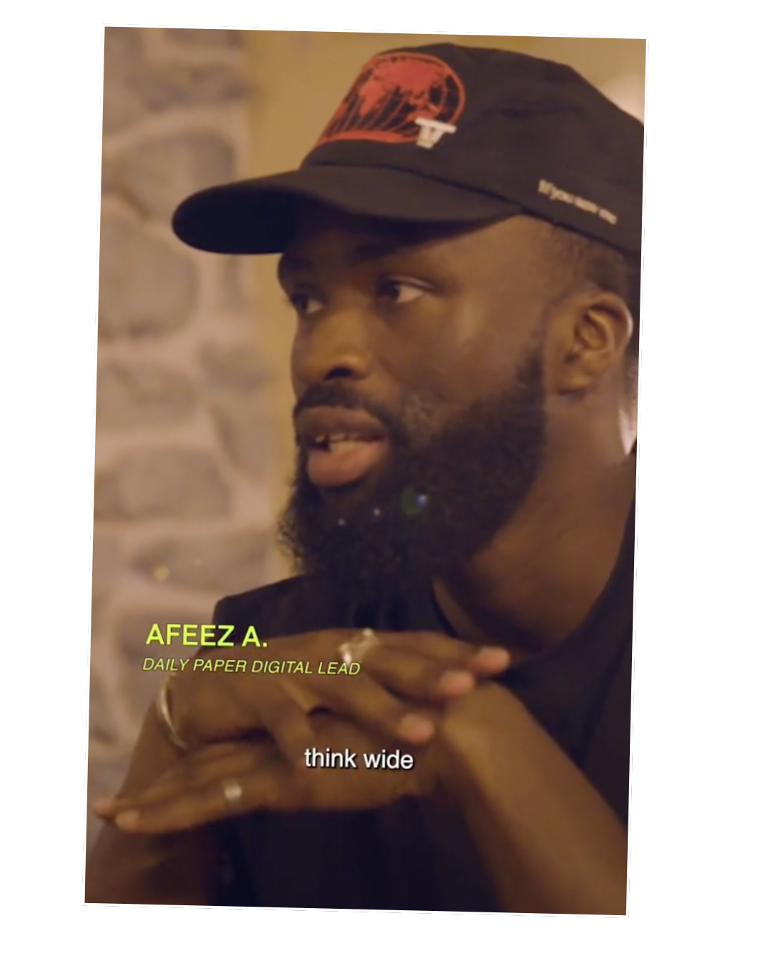 A man with a beard wearing a black cap with red graphics, sitting indoors against a stone wall, with text overlay that reads 'AFEZA A. DAILY PAPER DIGITAL LEAD think wide'.