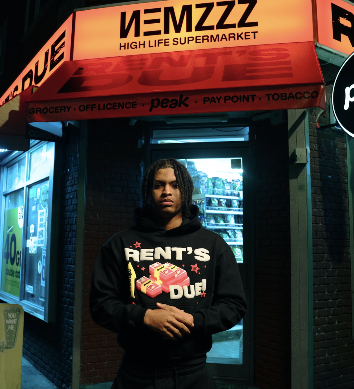 A young man with dreadlocks wearing a black hoodie with graphic text stands outside a convenience store at night. The store has a bright red sign with the words 'HIGH LIFE SUPERMARKET' and additional signage indicating it sells groceries, alcohol, pay points, and tobacco. The store appears to be in an urban area with illuminated vending machines and a brick facade.