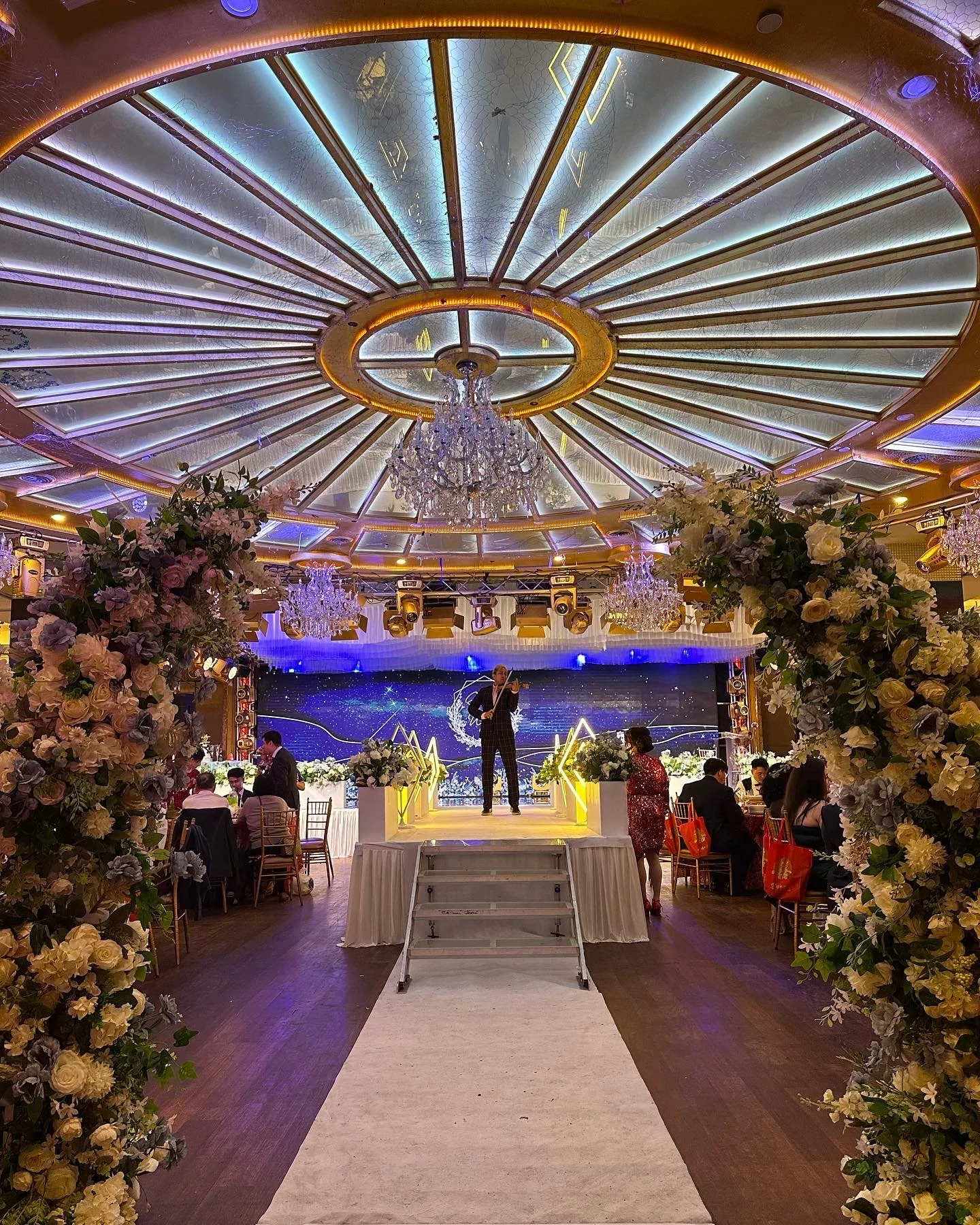 Elegant event venue with a floral archway, a stage with a performer playing a violin, and a decorated ceiling with chandeliers, lighting, and starry backdrop.