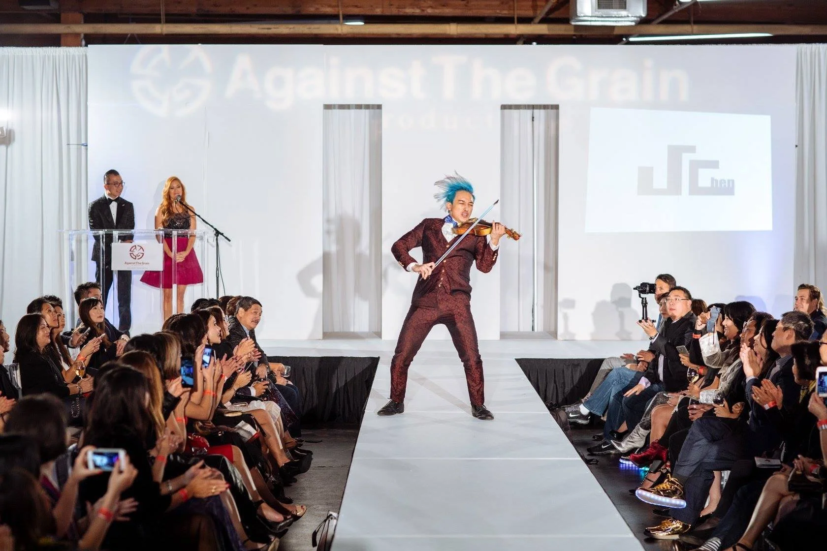 Performer with blue hair playing the violin on a runway at a fashion event, with an audience and speakers in the background.