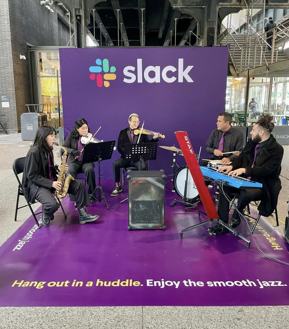A musical band performing on a purple stage with a large Slack logo in the background. The band includes musicians playing a saxophone, violin, drums, and keyboard.