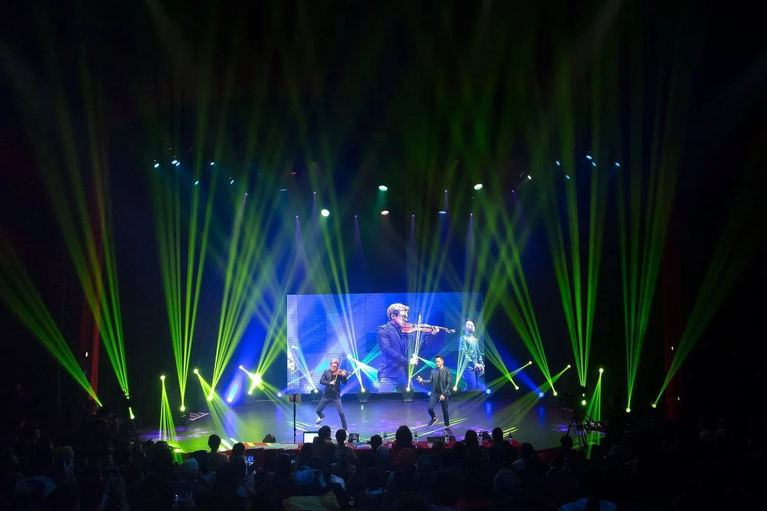 Stage with a large screen showing a violinist, surrounded by bright green and yellow stage lights, with performers dancing in front of an audience.