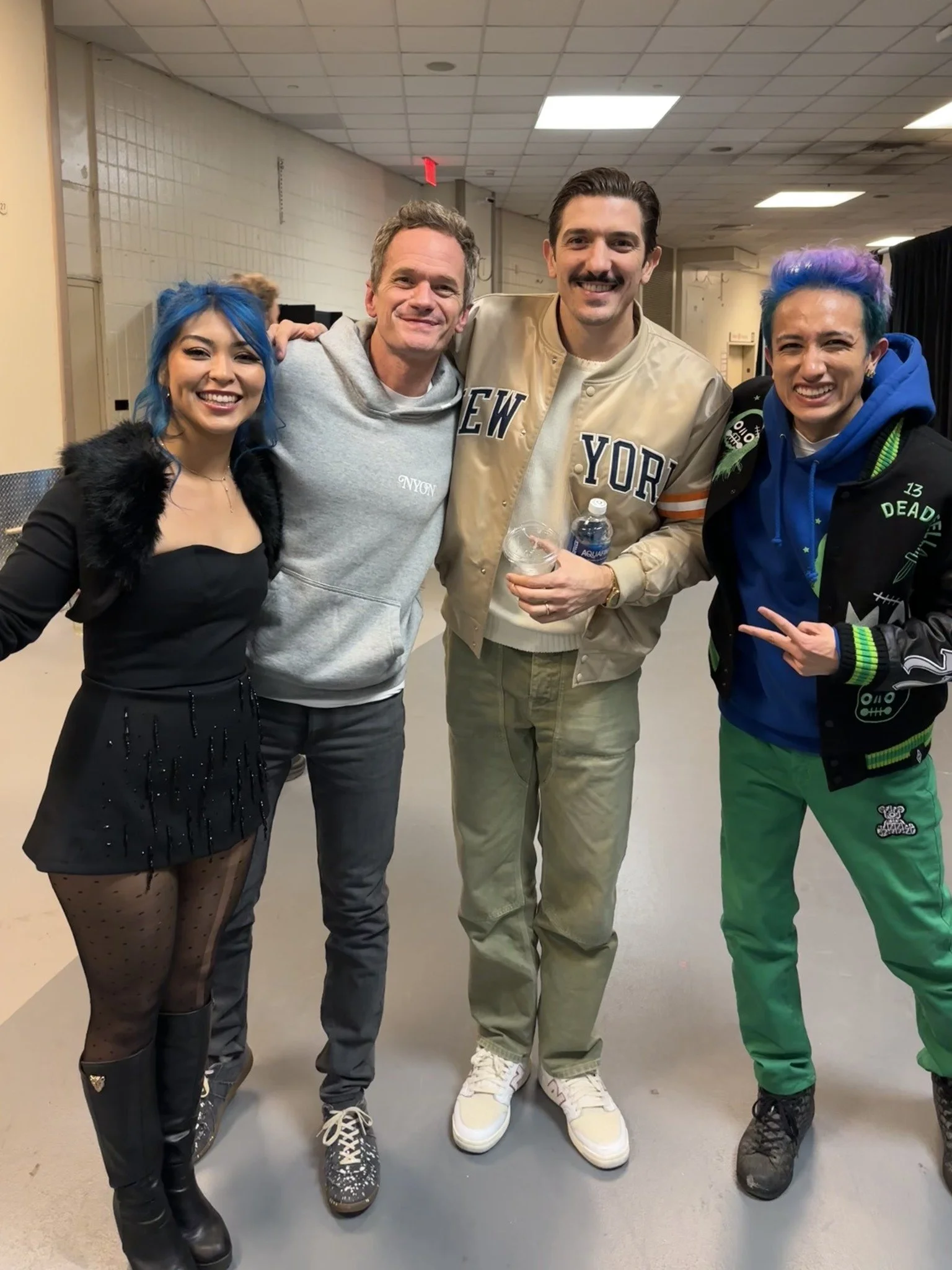 Four people standing indoors, smiling for the camera, with three appearing to be performers and one casually dressed, holding a water bottle and a plastic cup, in a hallway with ceiling lights.