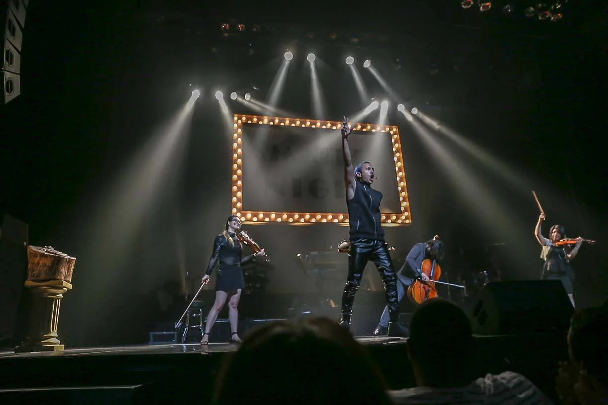 A musical performance on stage featuring a woman in black pants and a sleeveless top singing while raising her right hand, with four other musicians playing strings instruments underneath bright stage lights and a large illuminated rectangular frame