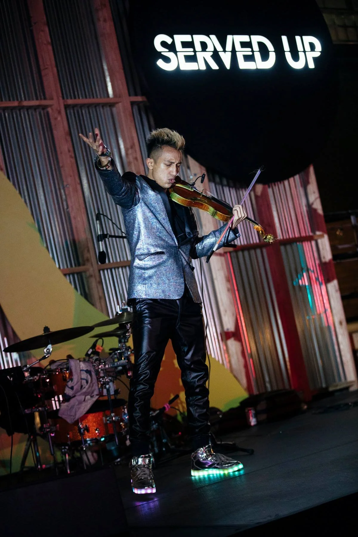A musician wearing a shiny silver jacket and black shiny pants, playing a violin on stage with a backdrop that reads 'Served Up' in large illuminated letters. The musician is standing on a stage with colorful lighting and a drum set visible behind hi