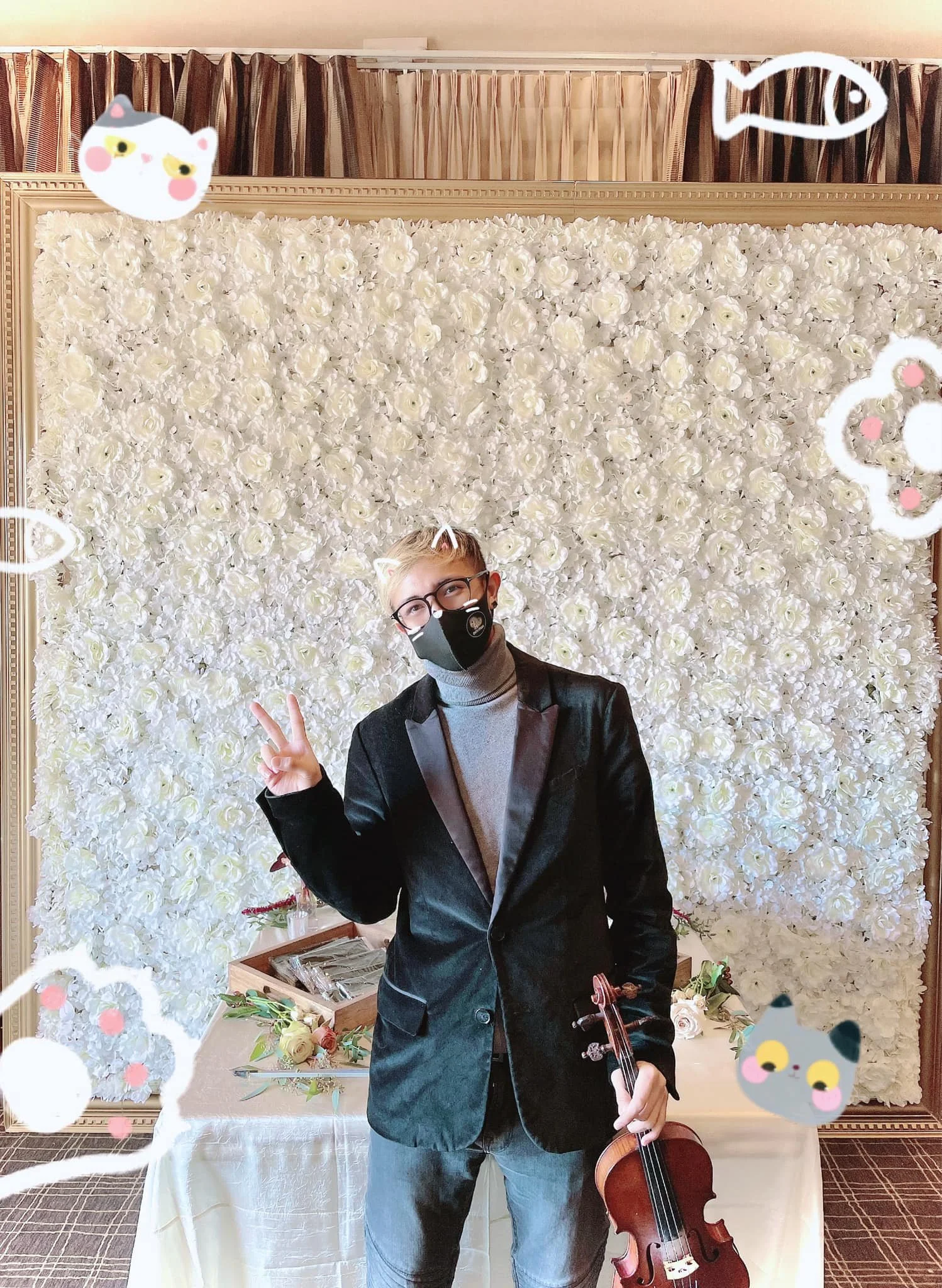 A man with blond hair, glasses, and a black face mask making a peace sign with his right hand, holding a violin in his left hand, standing in front of a flower wall decorated with cartoon animal and fish doodles.