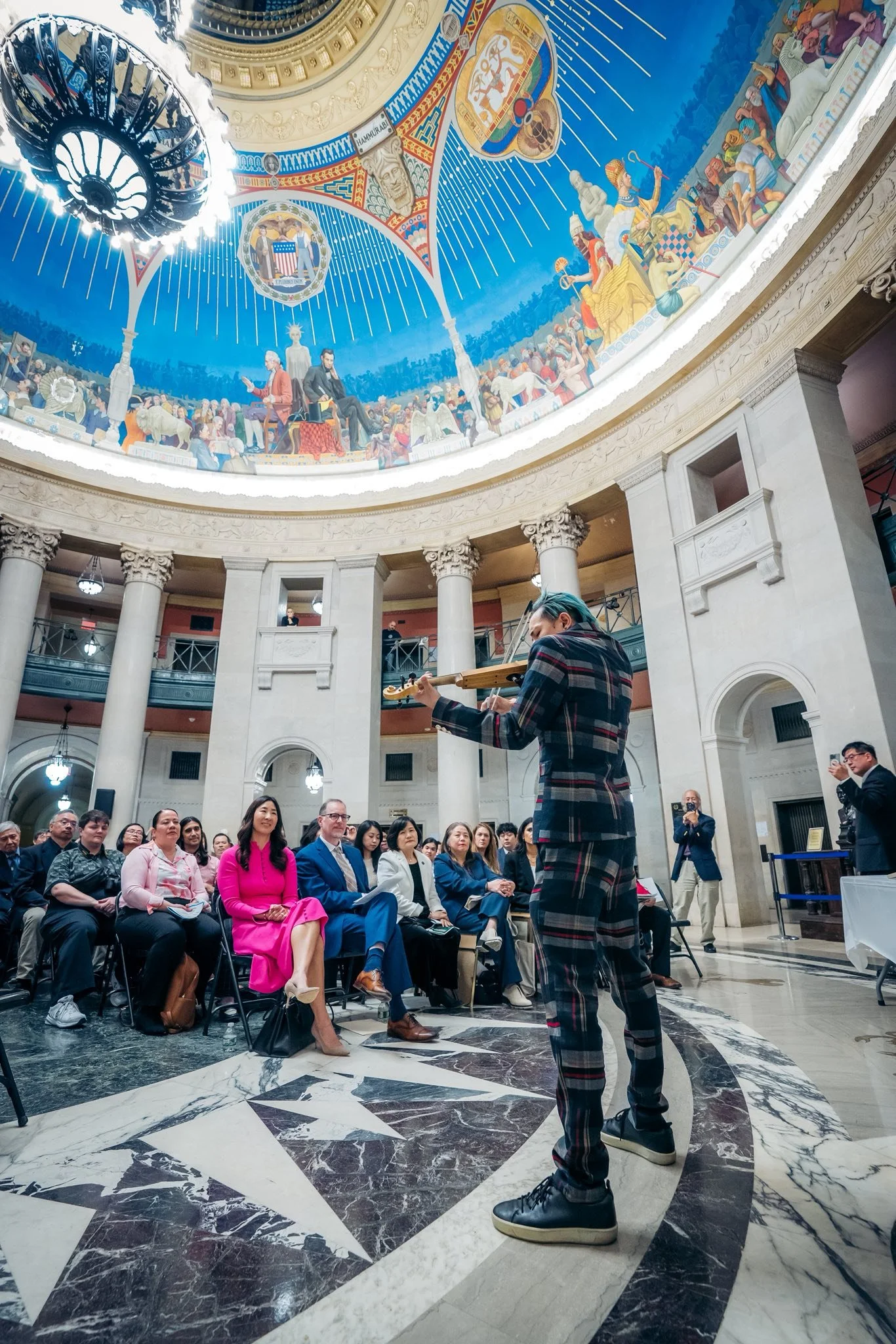 A musician playing a guitar at a musical performance in a grand hall with a domed ceiling. The audience is seated and watching attentively. The hall's architecture features tall columns and a marble floor. The ceiling displays a large colorful mural