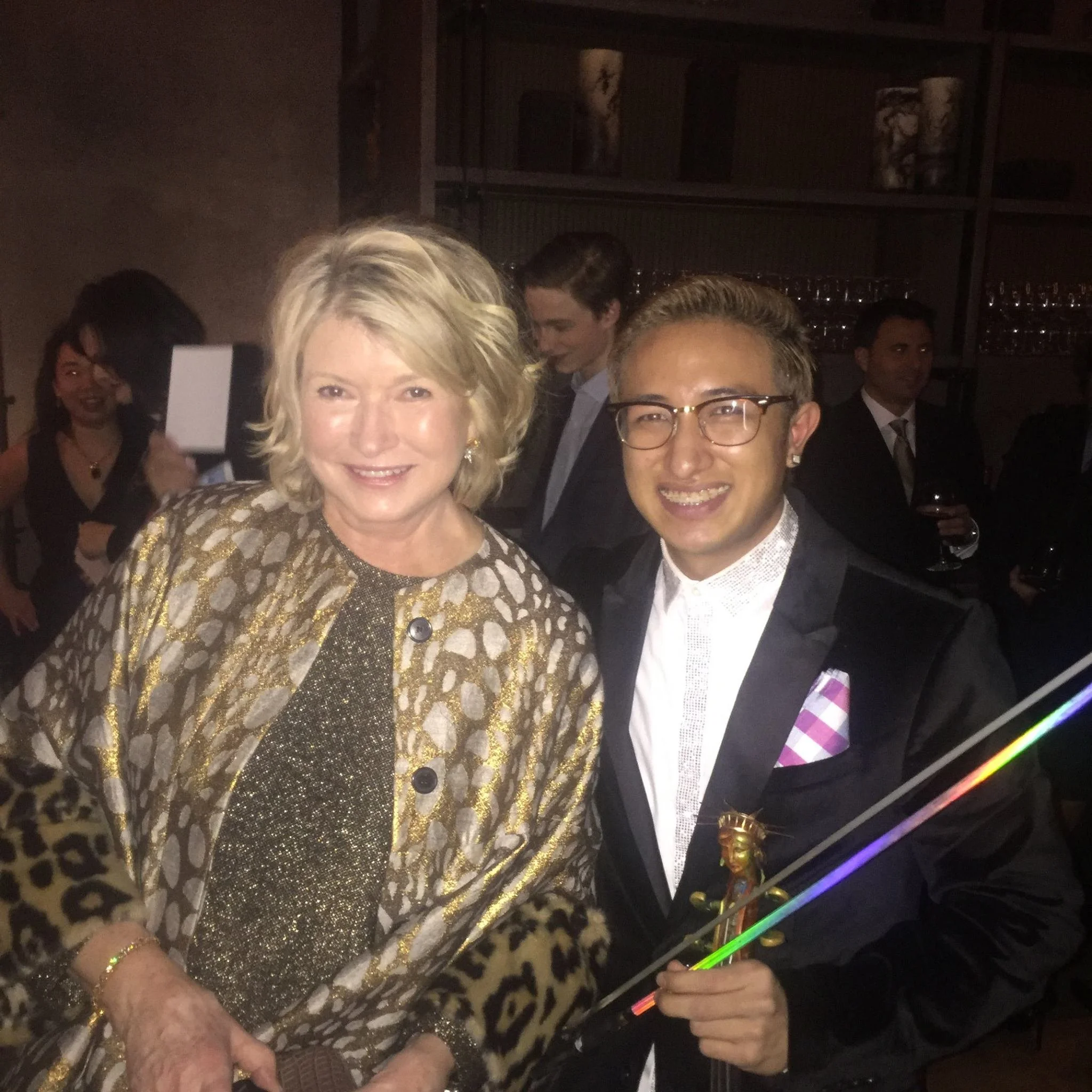 A woman with blonde hair wearing a gold and animal print coat standing next to a man with glasses, brown hair, and a black suit at a social event, with other people in the background holding drinks.