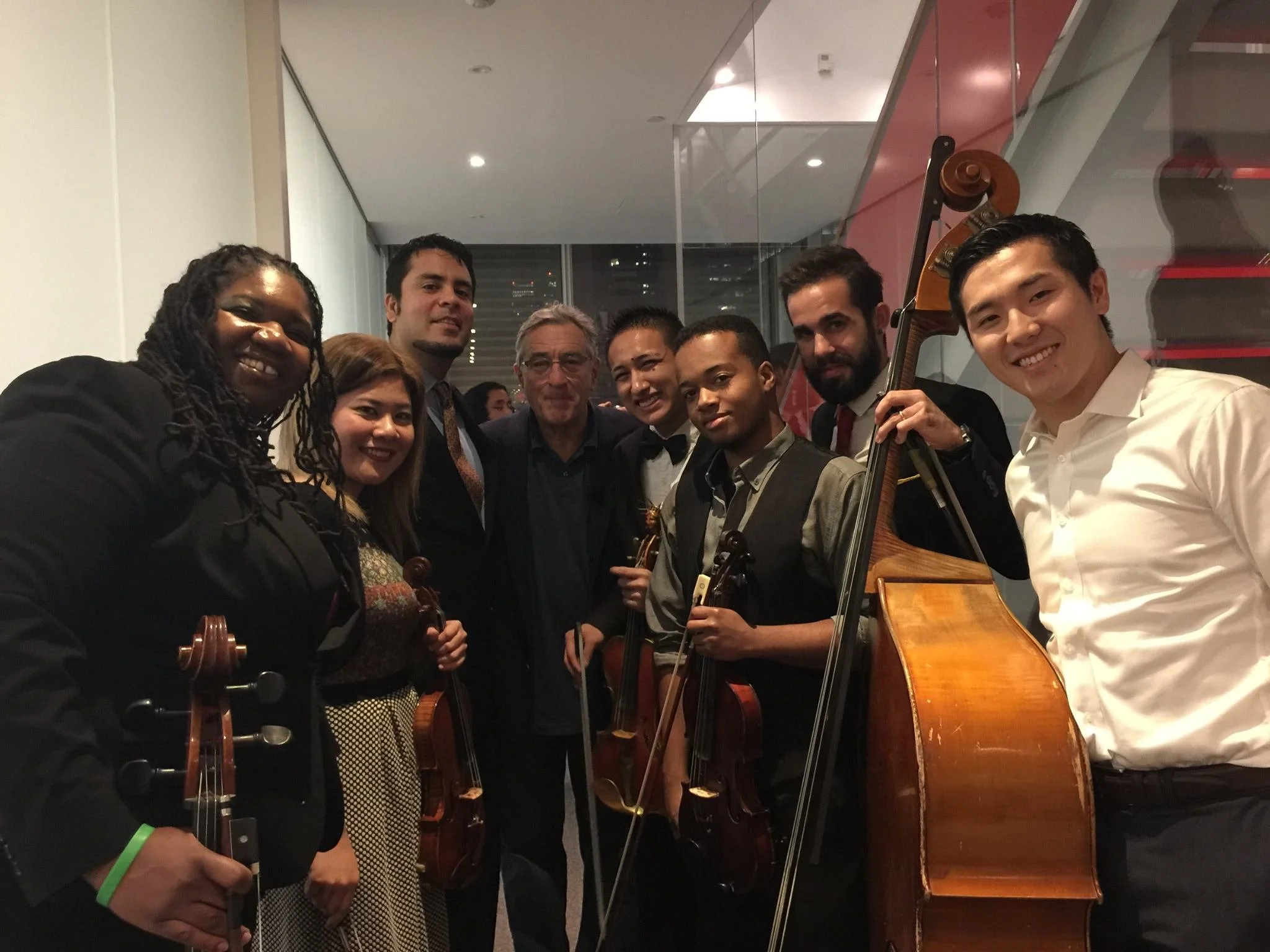 Group of nine diverse musicians holding violins and a double bass, smiling at the camera indoors.