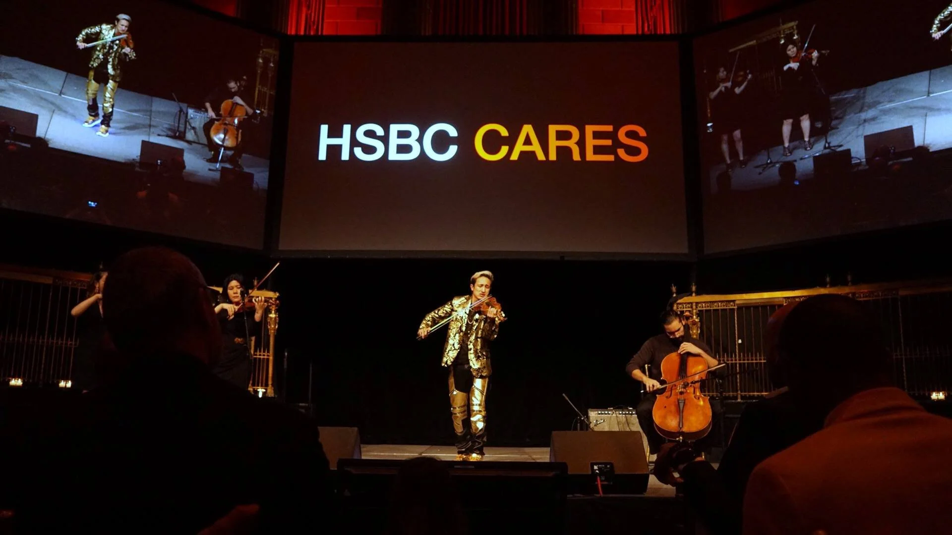 A stage with musicians performing, including a violinist and cellist, under a large screen displaying 'HSBC CARES', with smaller screens showing the performance from different angles.