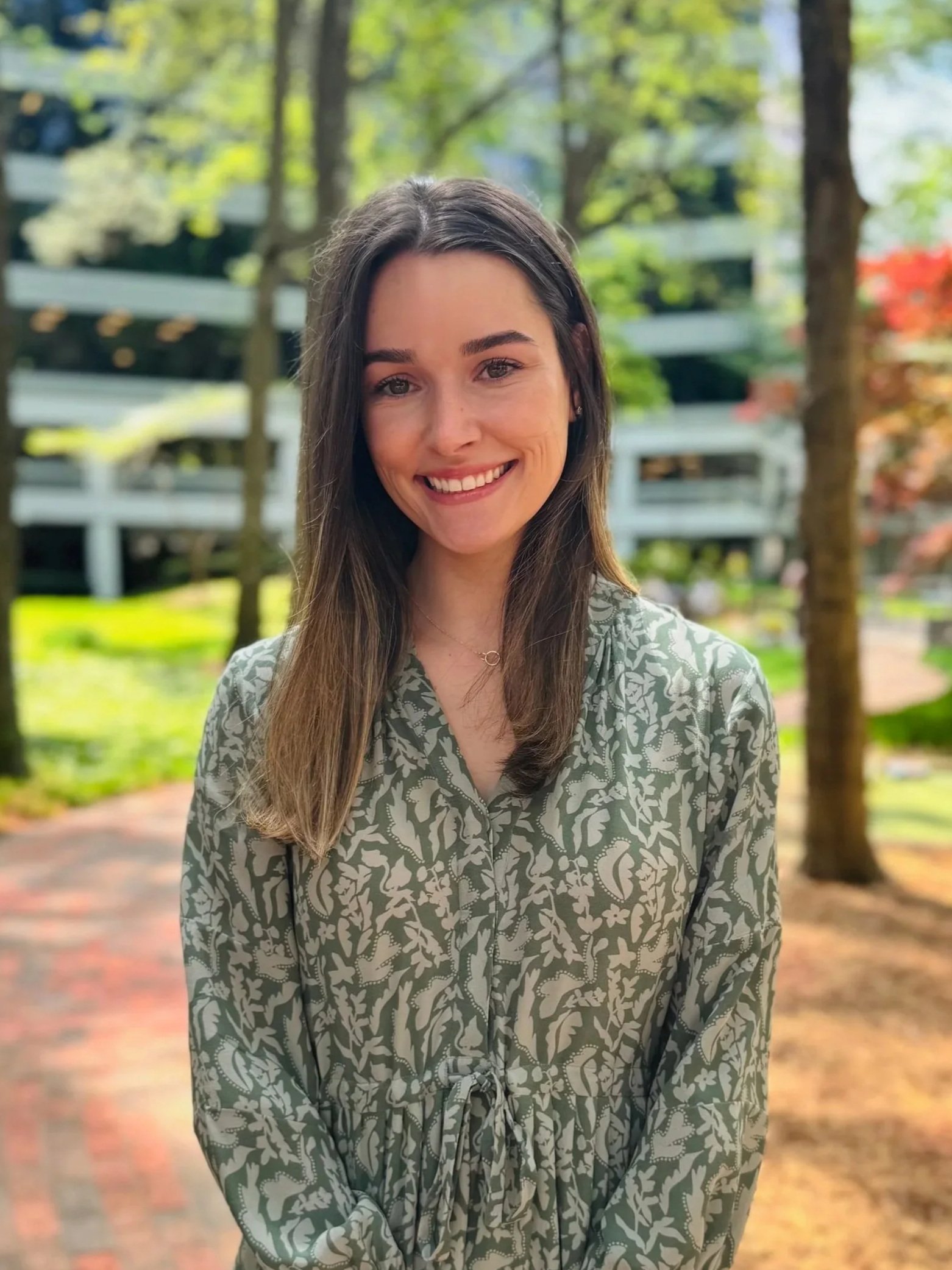 A young woman with shoulder-length brown hair and a big smile, wearing a blue blouse, outdoors with trees and greenery in the background.