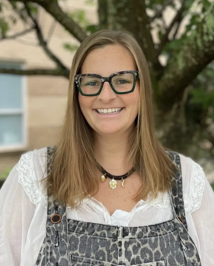 Smiling woman with long light brown hair, wearing black glasses, a white blouse with lace details, and a leopard print overall dress, standing outdoors with a tree and house in the background.