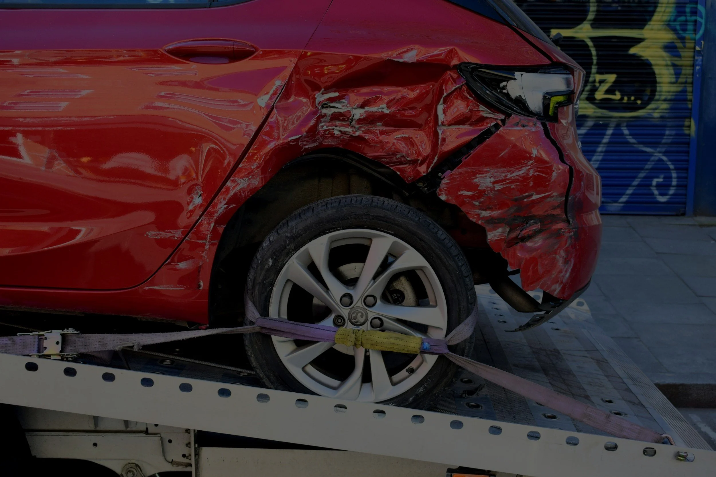 Coche rojo con daños por un accidente, asegurado en una plataforma de remolque con cinturones de seguridad.