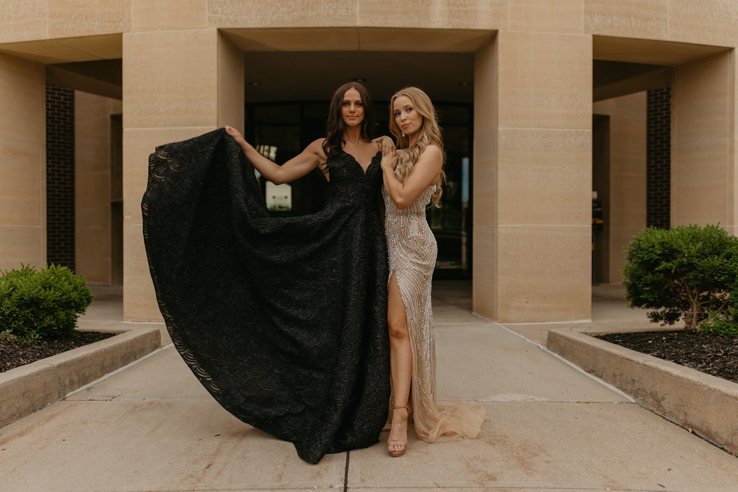 Two women in glamorous dresses standing on the steps of a historic building, posing and wearing sunglasses.