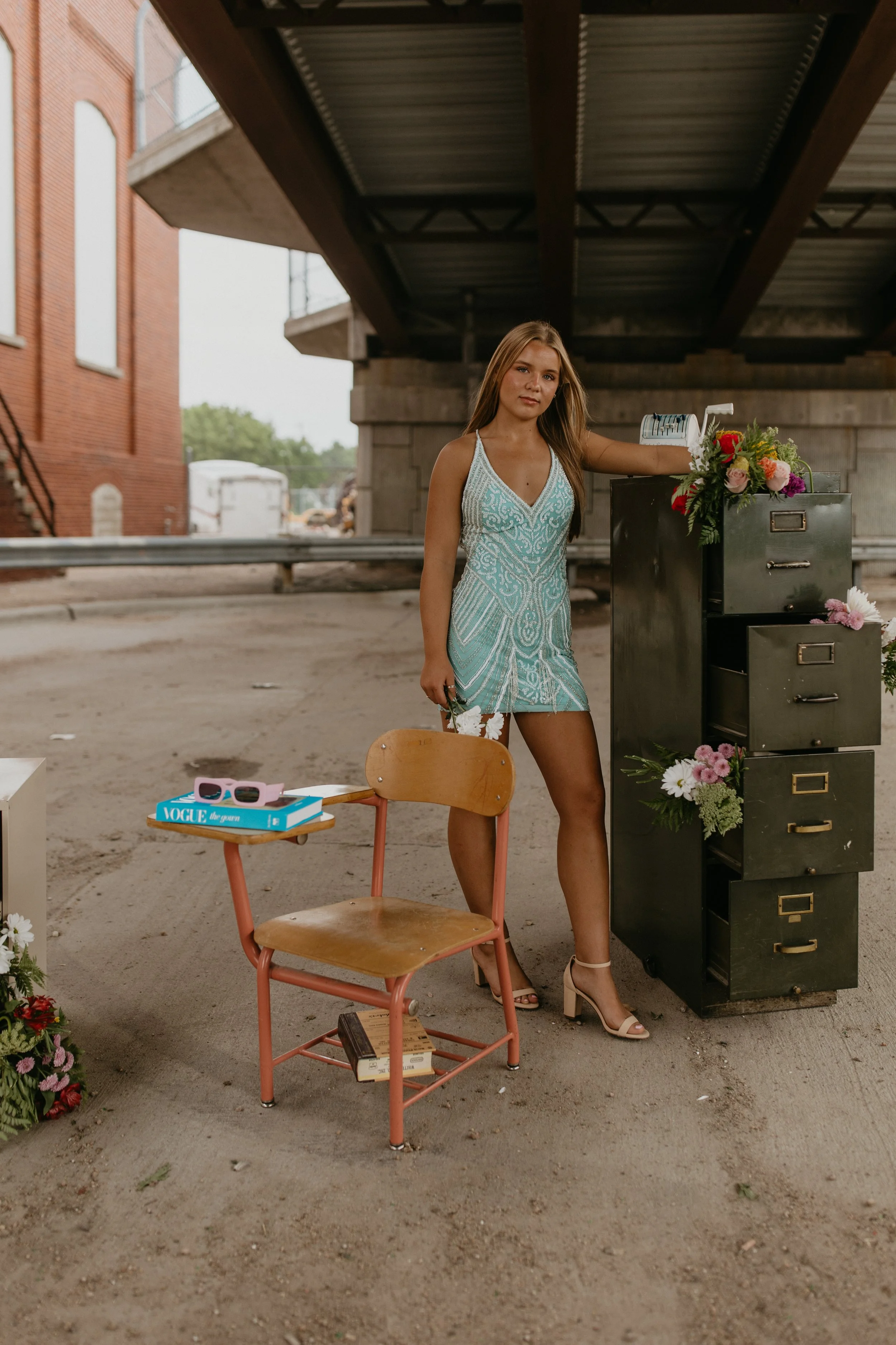 A young woman in a patterned dress and high heels standing beside a black filing cabinet with flowers and a paper shredder on top, outdoors under a bridge with a brick building in the background.