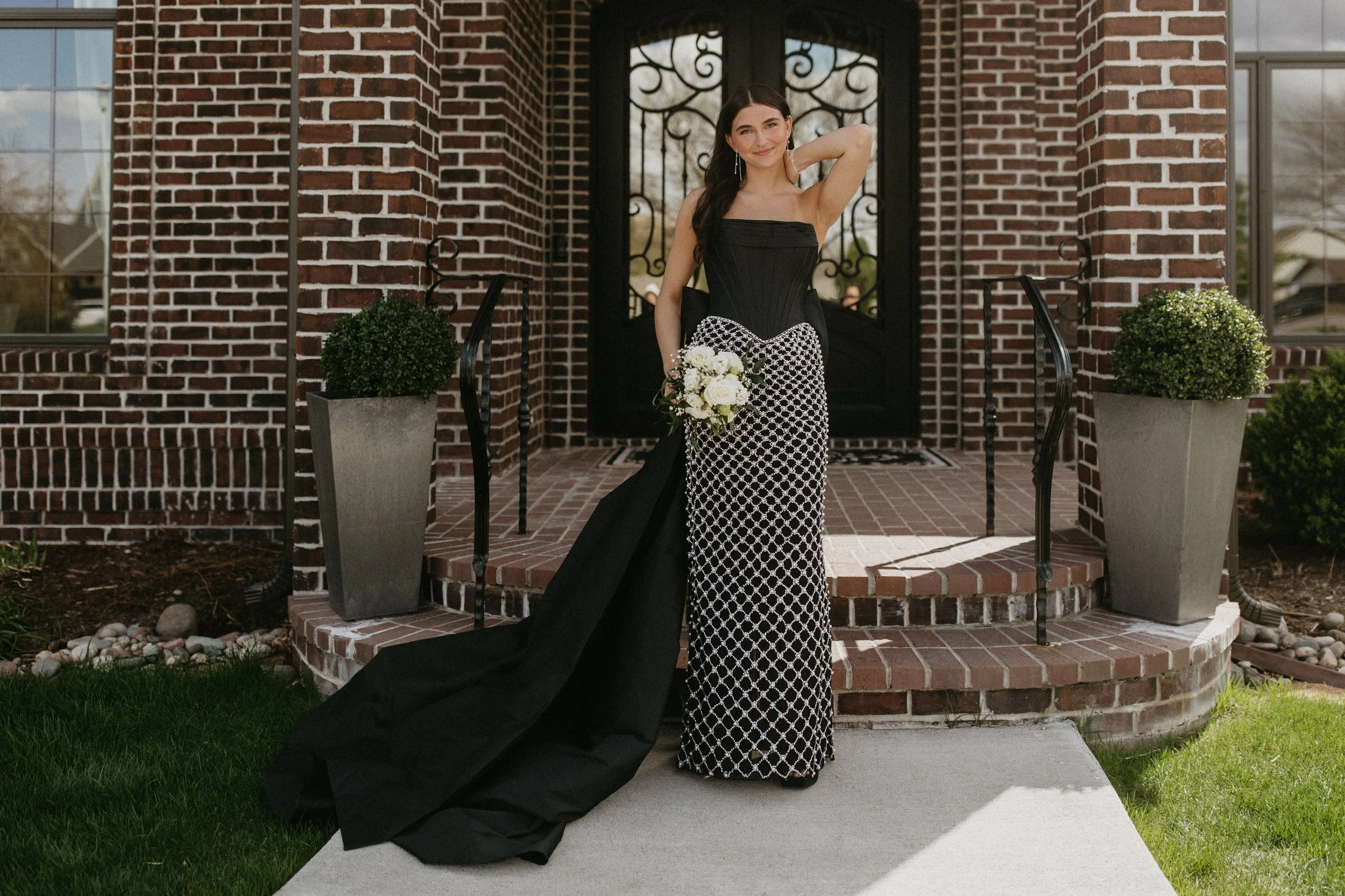 A young woman in a formal black and white gown standing on a porch in front of a brick house, holding a bouquet of white flowers, smiling, with her right hand raised to her head.