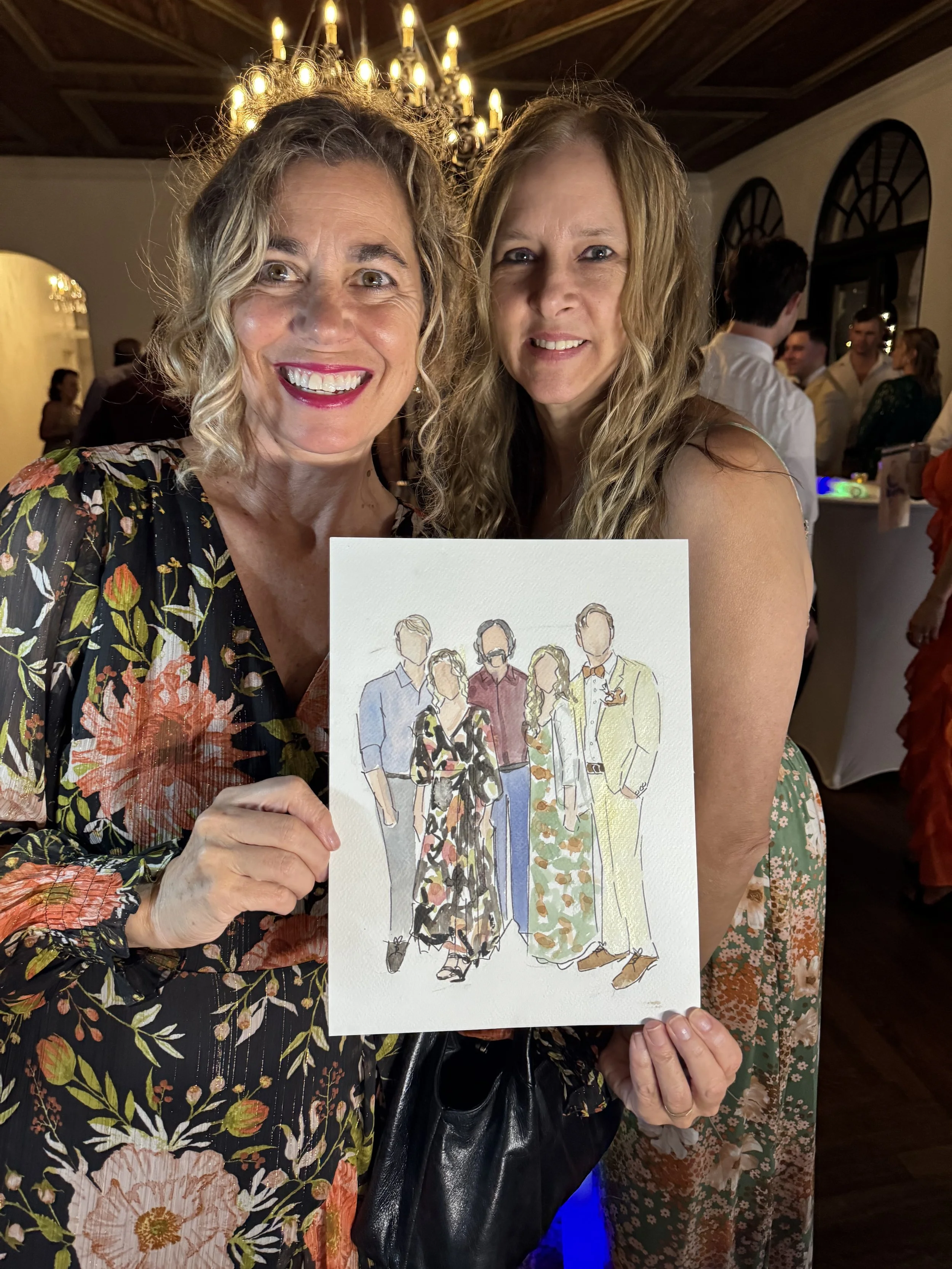 Two women holding a drawing of a group of people at a party or celebration, smiling at the camera, with a chandelier overhead and a crowd in the background.