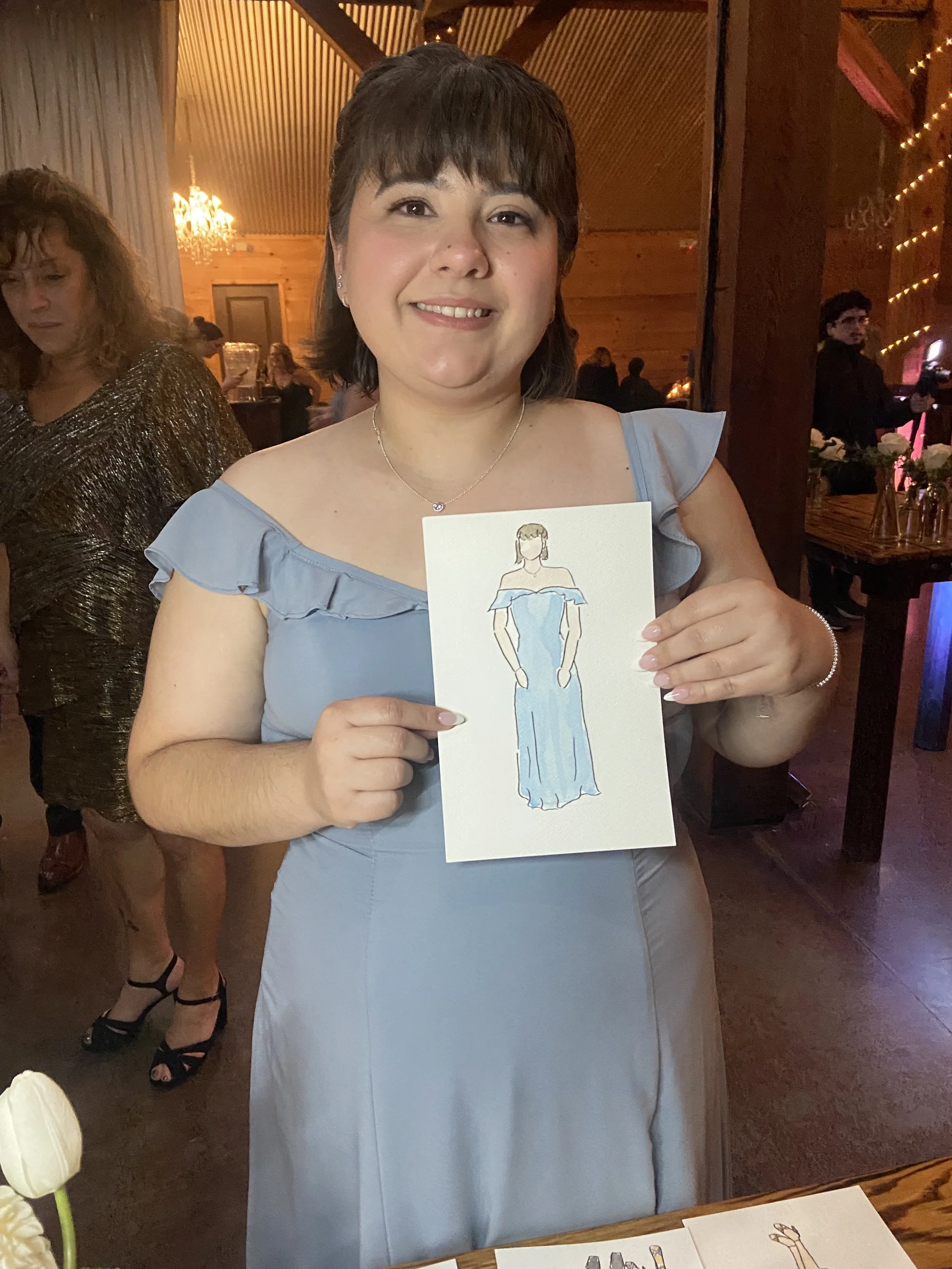 Woman in a blue dress holding a drawing of a similar off-the-shoulder blue gown at a celebration or party.