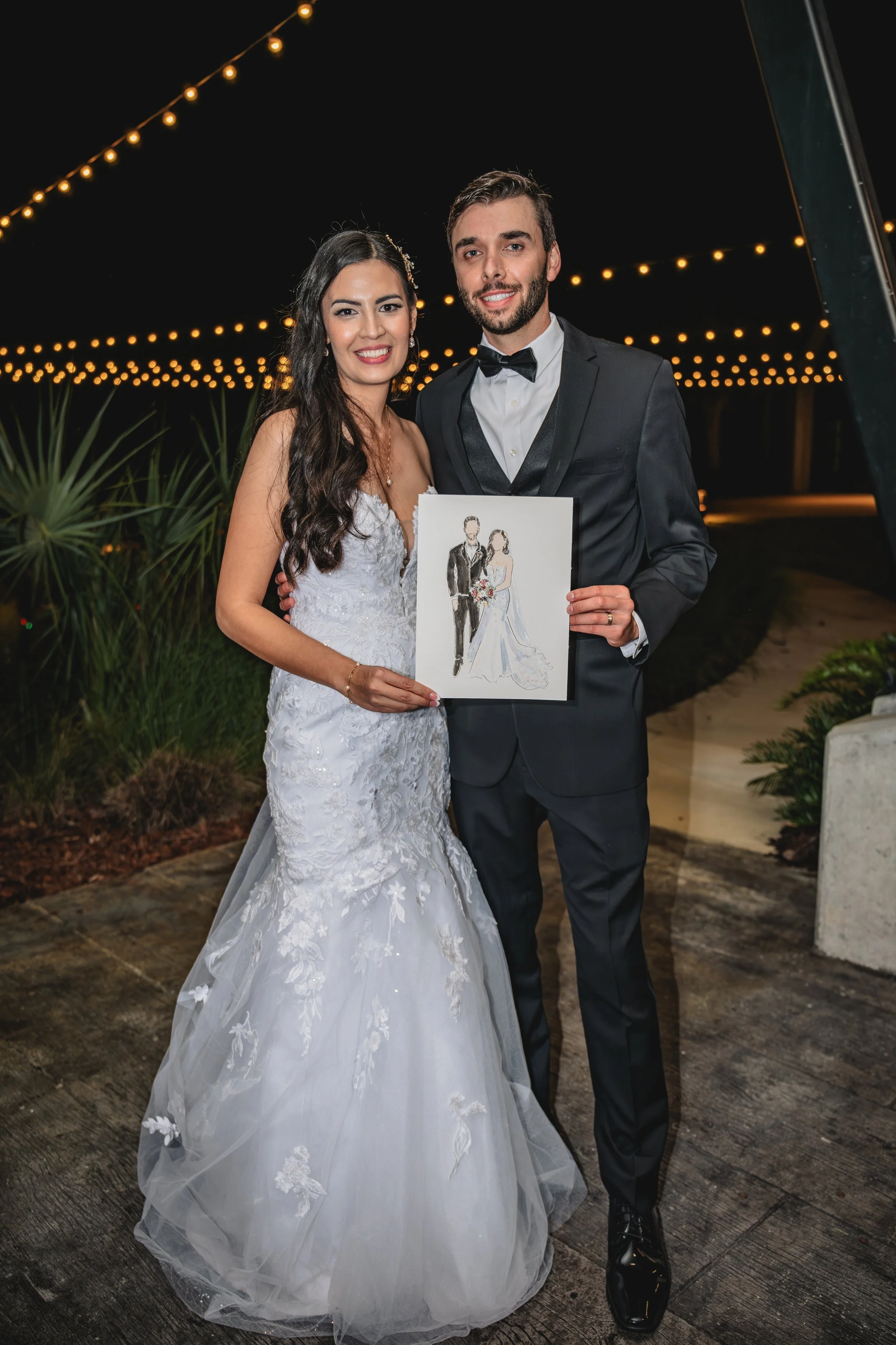 Bride and groom holding a drawing of themselves in wedding attire at night under string lights.