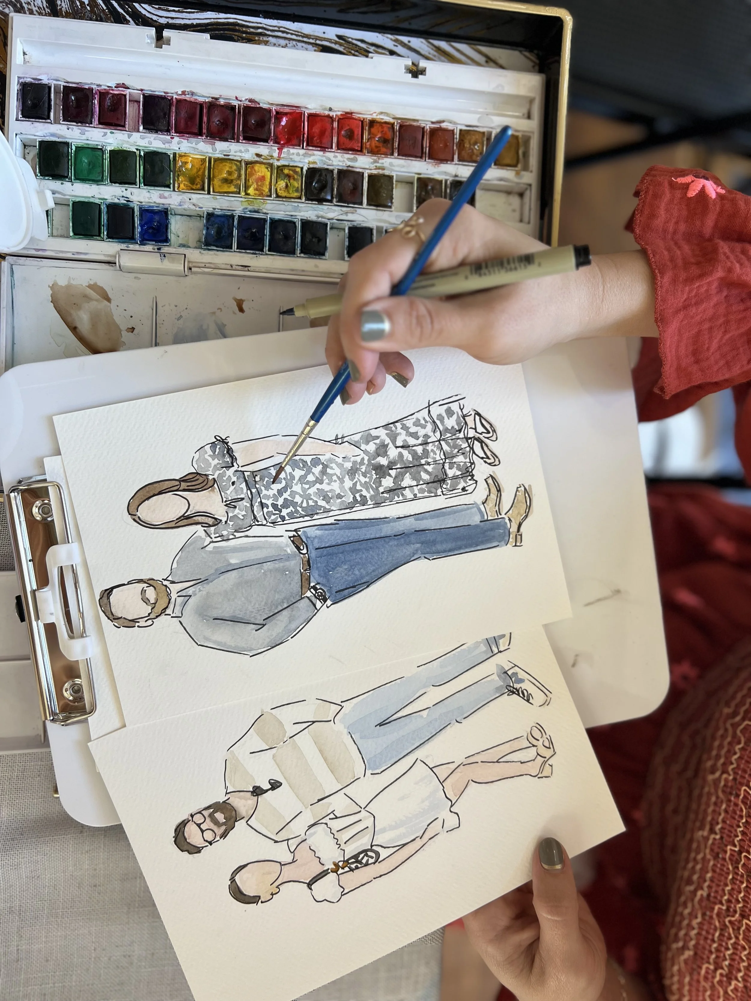 Artist painting fashion sketches of a man and a woman using watercolors, with a watercolor palette nearby.