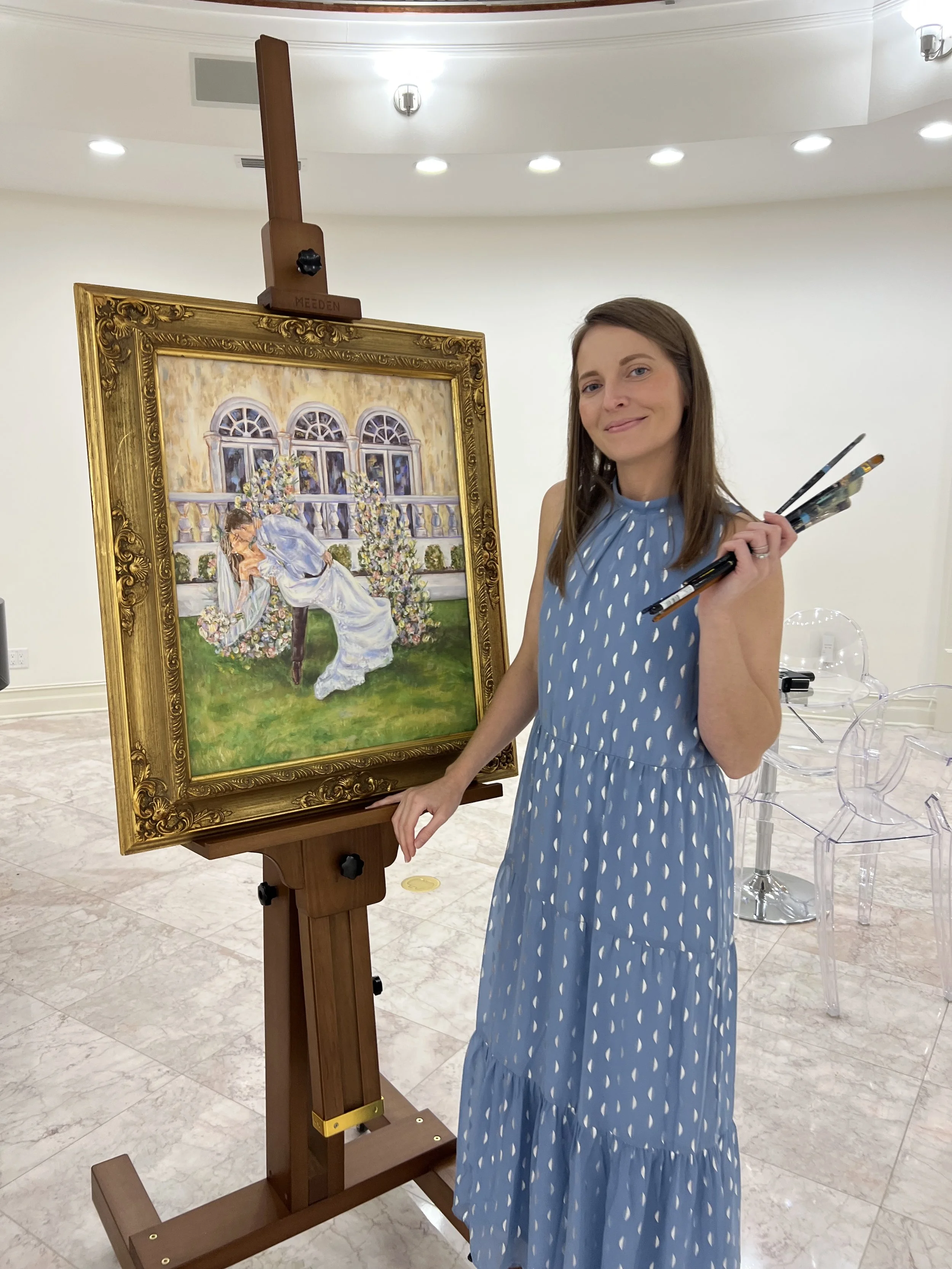 Brittany standing next to an easel with a framed painting of a woman lying on the grass near a building with arched windows and flowering bushes.