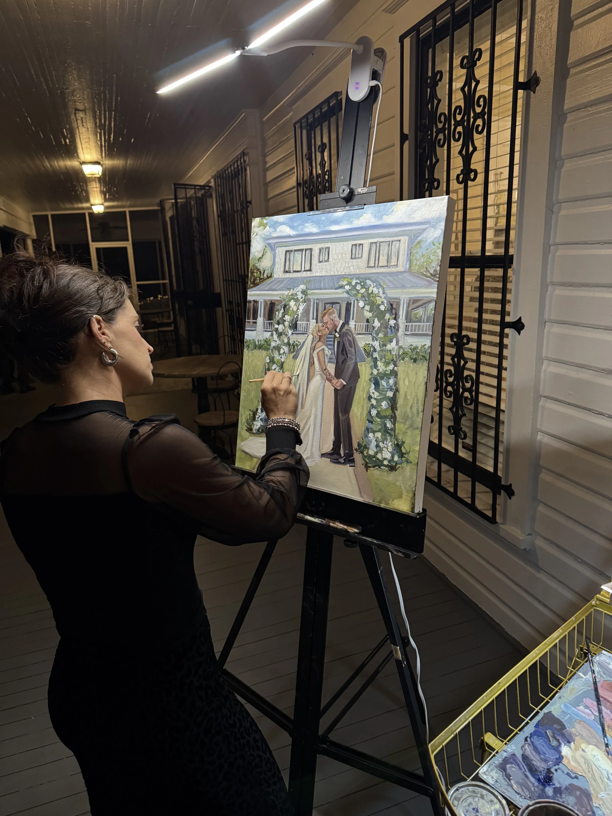 Holly painting wedding portrait on canvas, depicting a couple at an outdoor wedding ceremony with an arch and a house in the background. She is standing indoors at an easel illuminated by an overhead light.