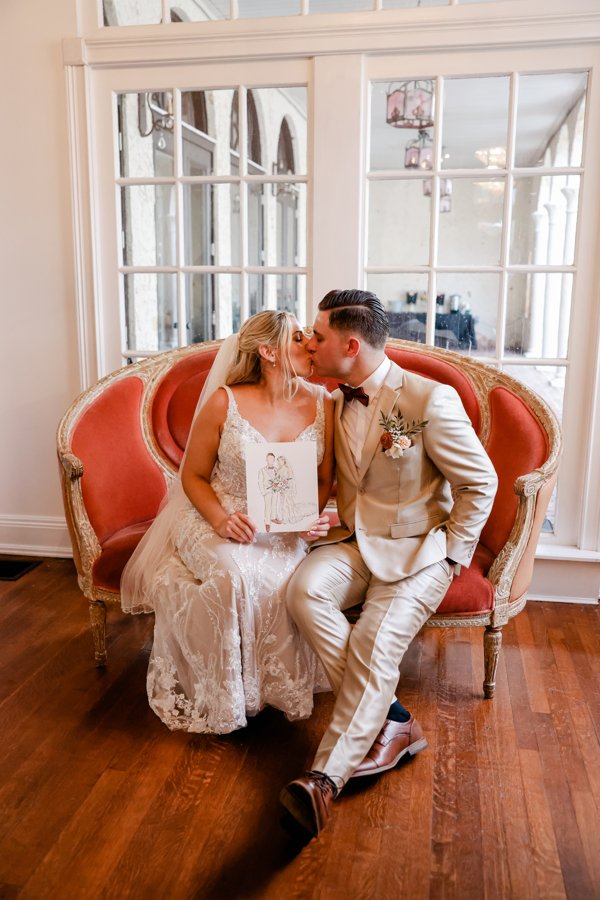 A bride and groom sitting on a vintage pink sofa, sharing a kiss at their wedding. The bride is holding an illustration of a wedding couple. The setting has large windows and wooden flooring.