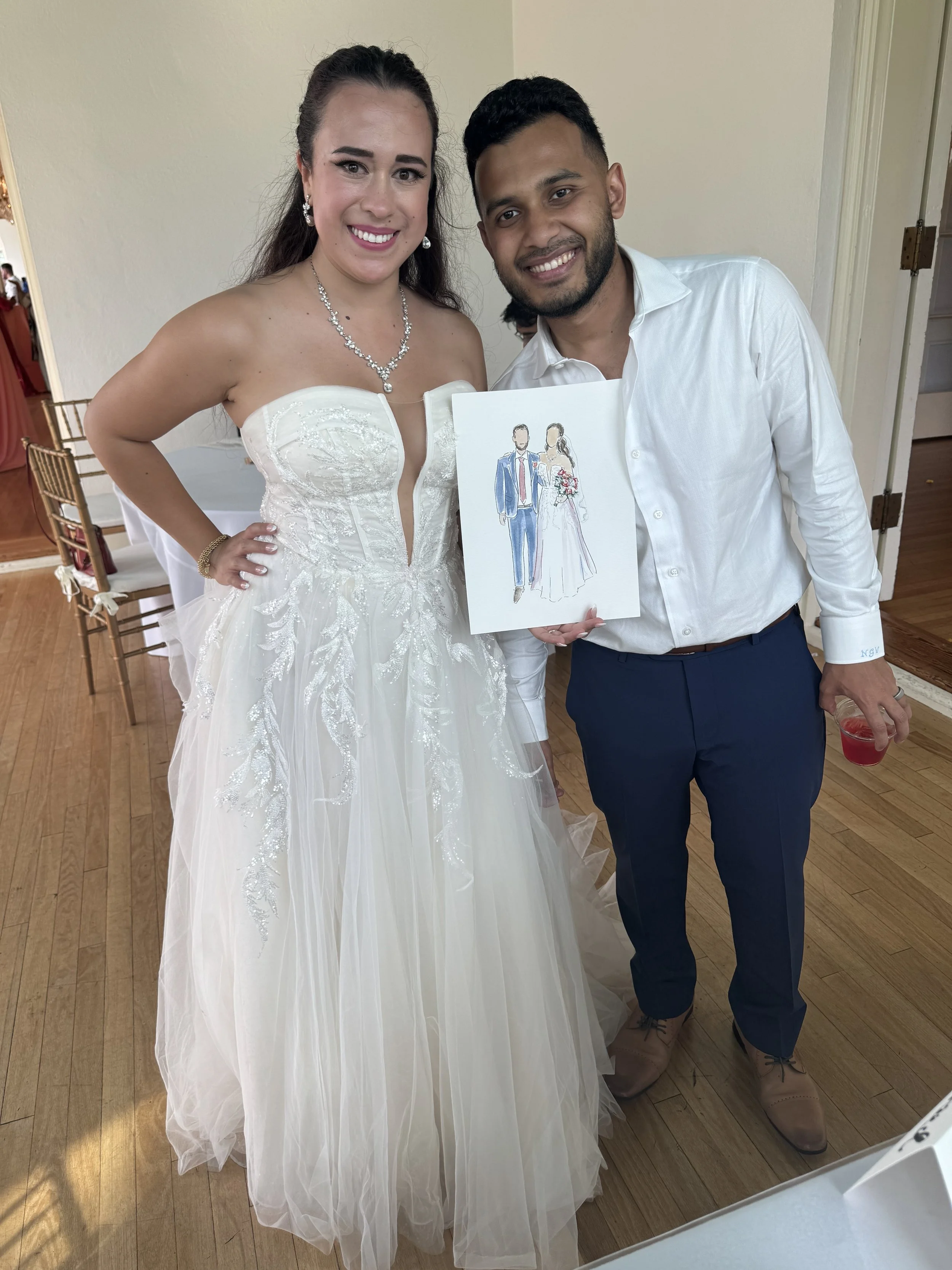 A smiling bride in a white wedding dress with embroidery and a necklace standing beside a man in a white shirt and blue pants. They are holding a drawing of a couple dressed in wedding attire. The background shows wooden flooring and chairs.